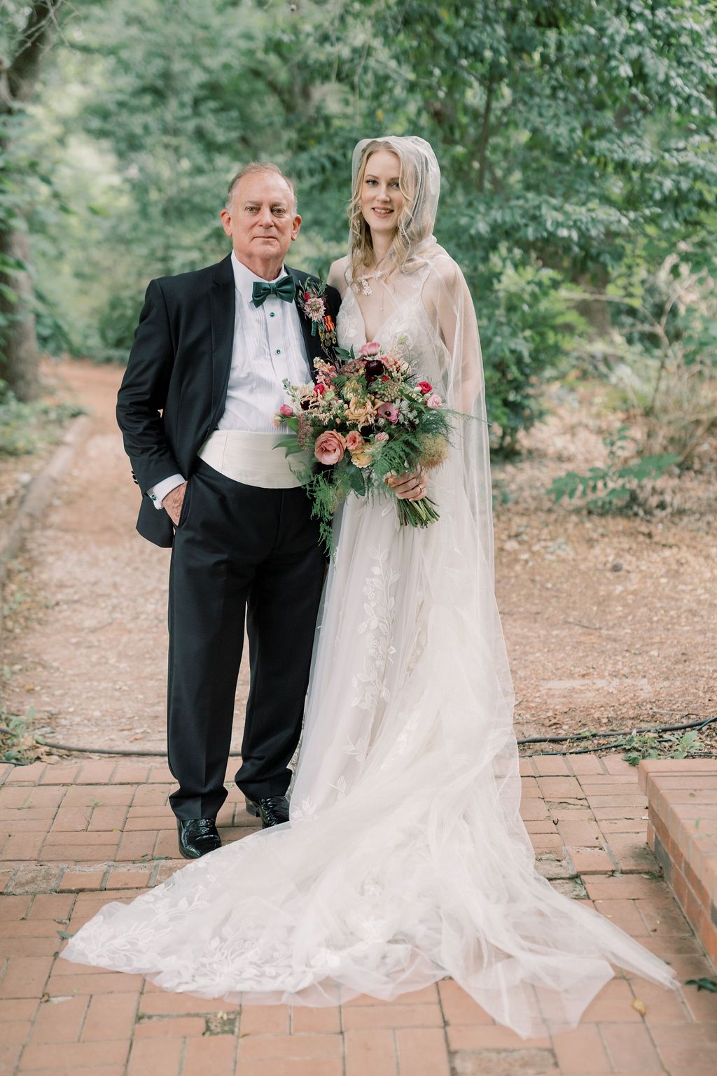 Bride in lace gown with veil holds bouquet beside man in tuxedo on brick path surrounded by lush greenery.