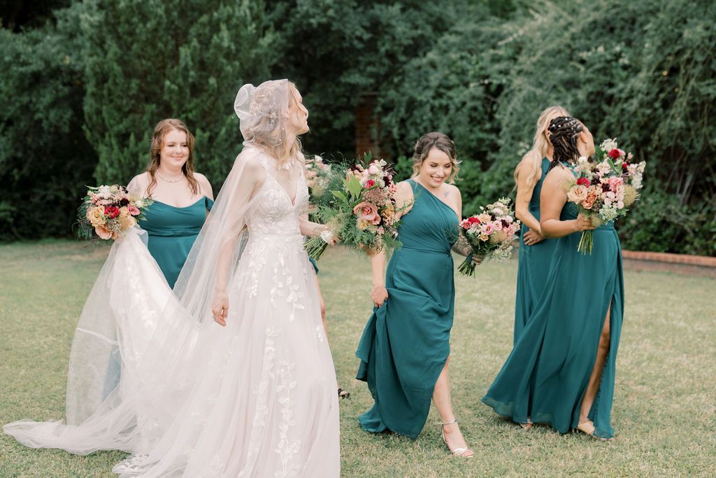 Bride in white gown walks with three bridesmaids in teal dresses holding colorful bouquets on a green lawn.