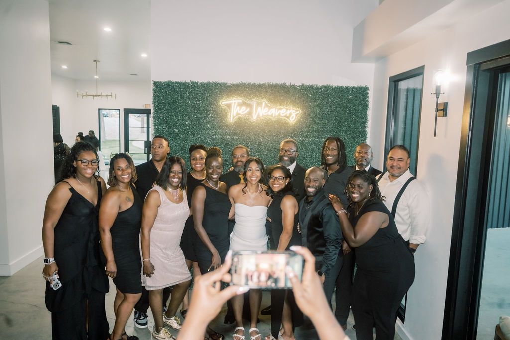 Group of elegantly dressed people posing together indoors near a green hedge wall with neon sign.