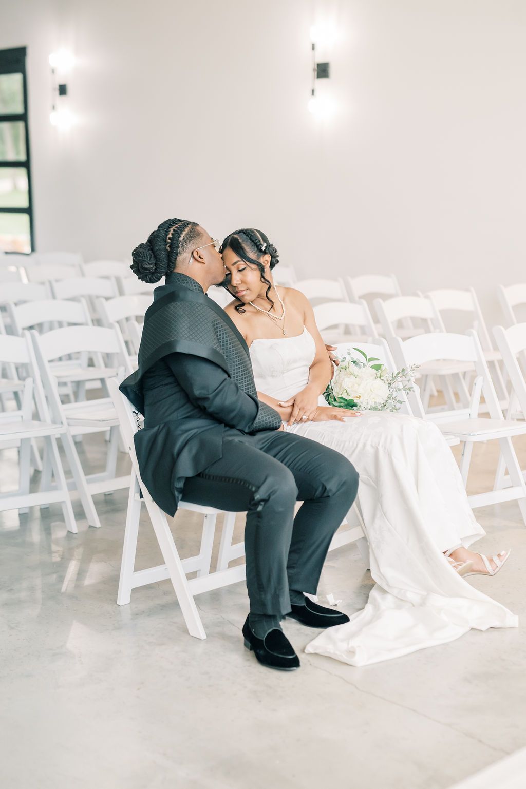 A bride and groom share an intimate moment seated among white chairs in a bright wedding venue.