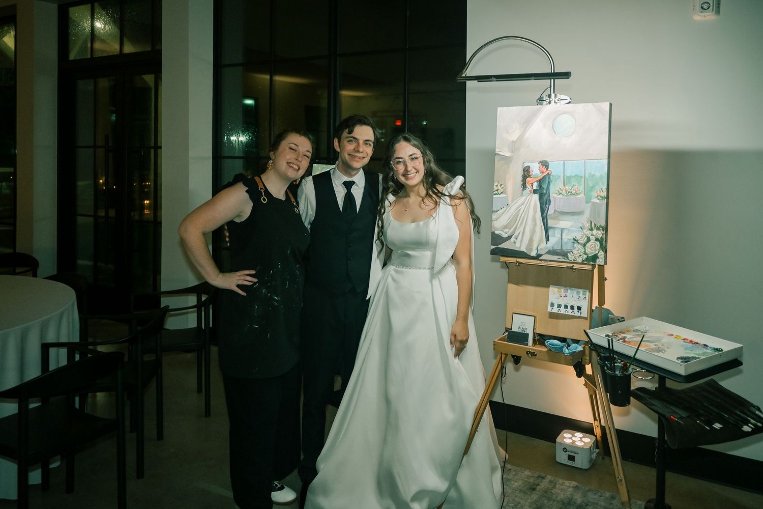 Bride, groom, and live wedding painter posing together beside a completed wedding portrait painting.