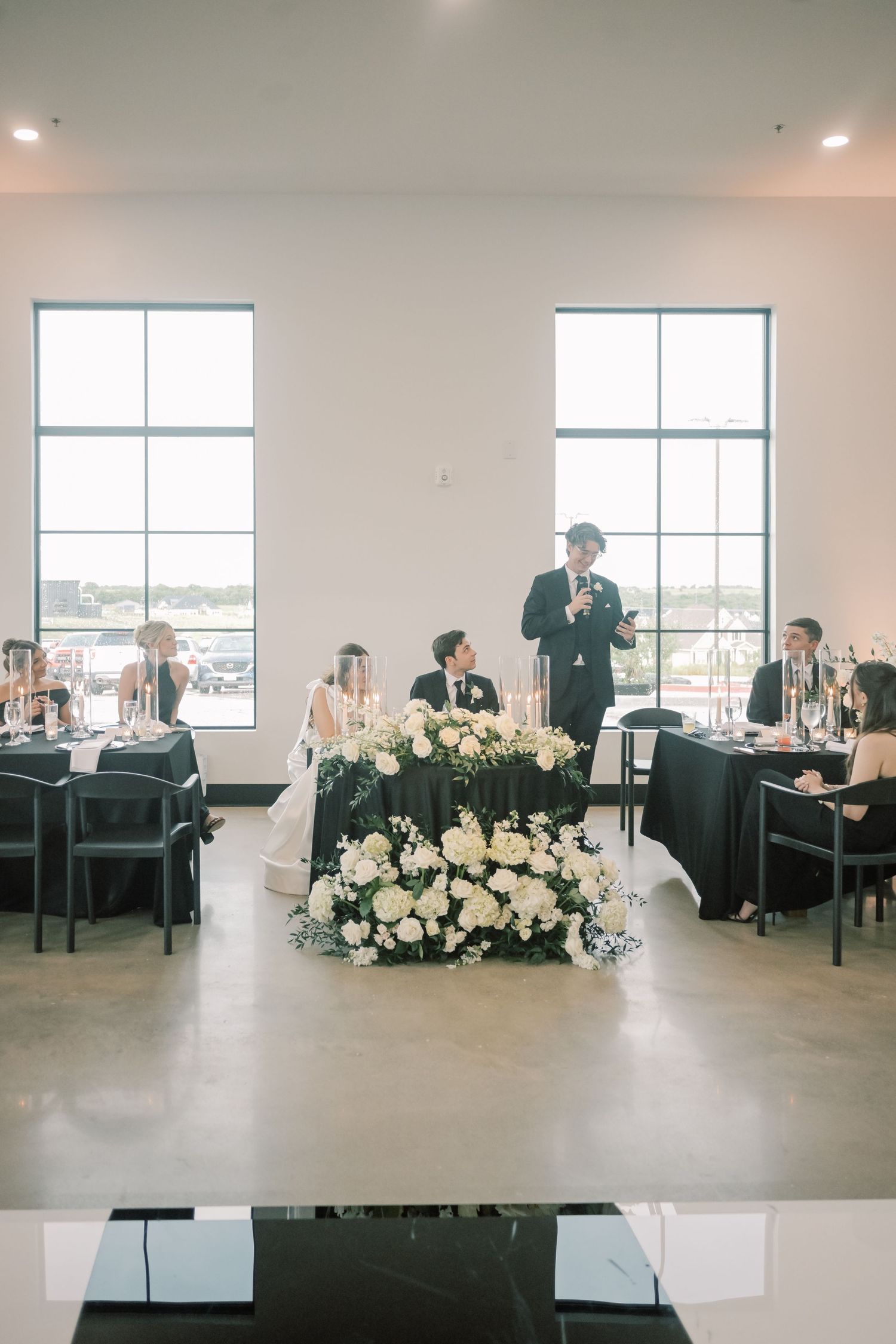 Newlyweds at sweetheart table adorned with white floral arrangement as groomsman delivers heartfelt toast.