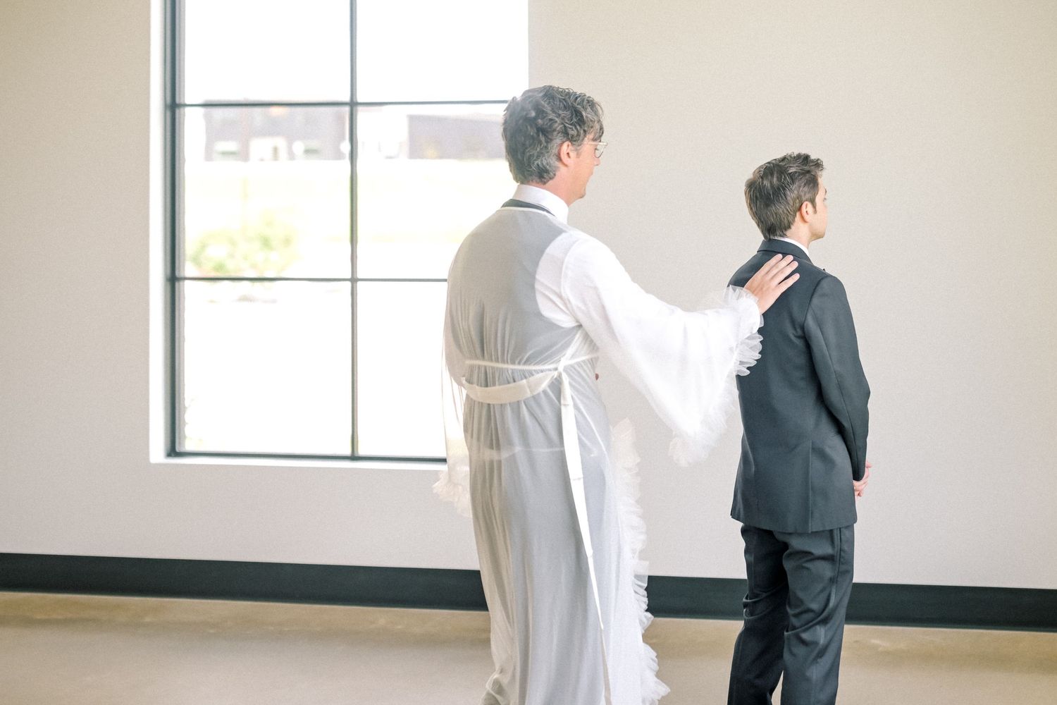 Bride in white wedding gown places hand on groom's shoulder during an intimate first look moment.