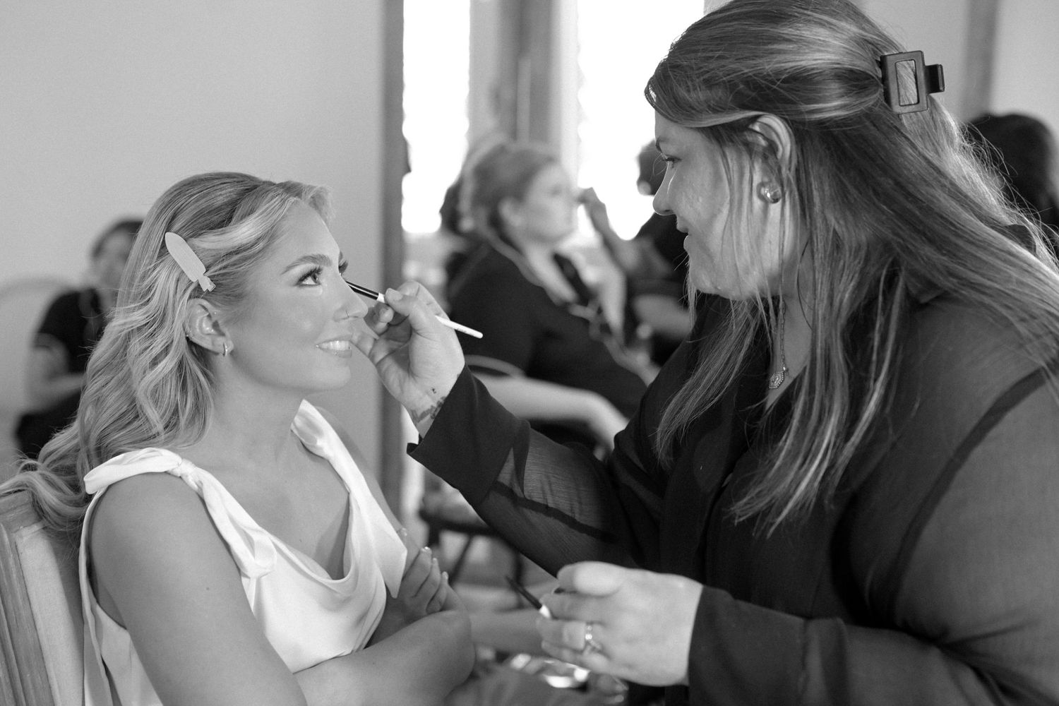 A makeup artist applies eyeshadow to a smiling blonde bride during wedding day preparations.