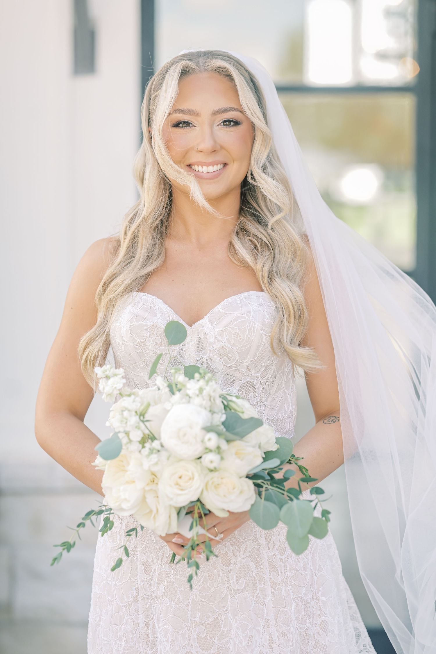 Radiant blonde bride smiles in strapless lace gown holding white roses and eucalyptus bouquet.