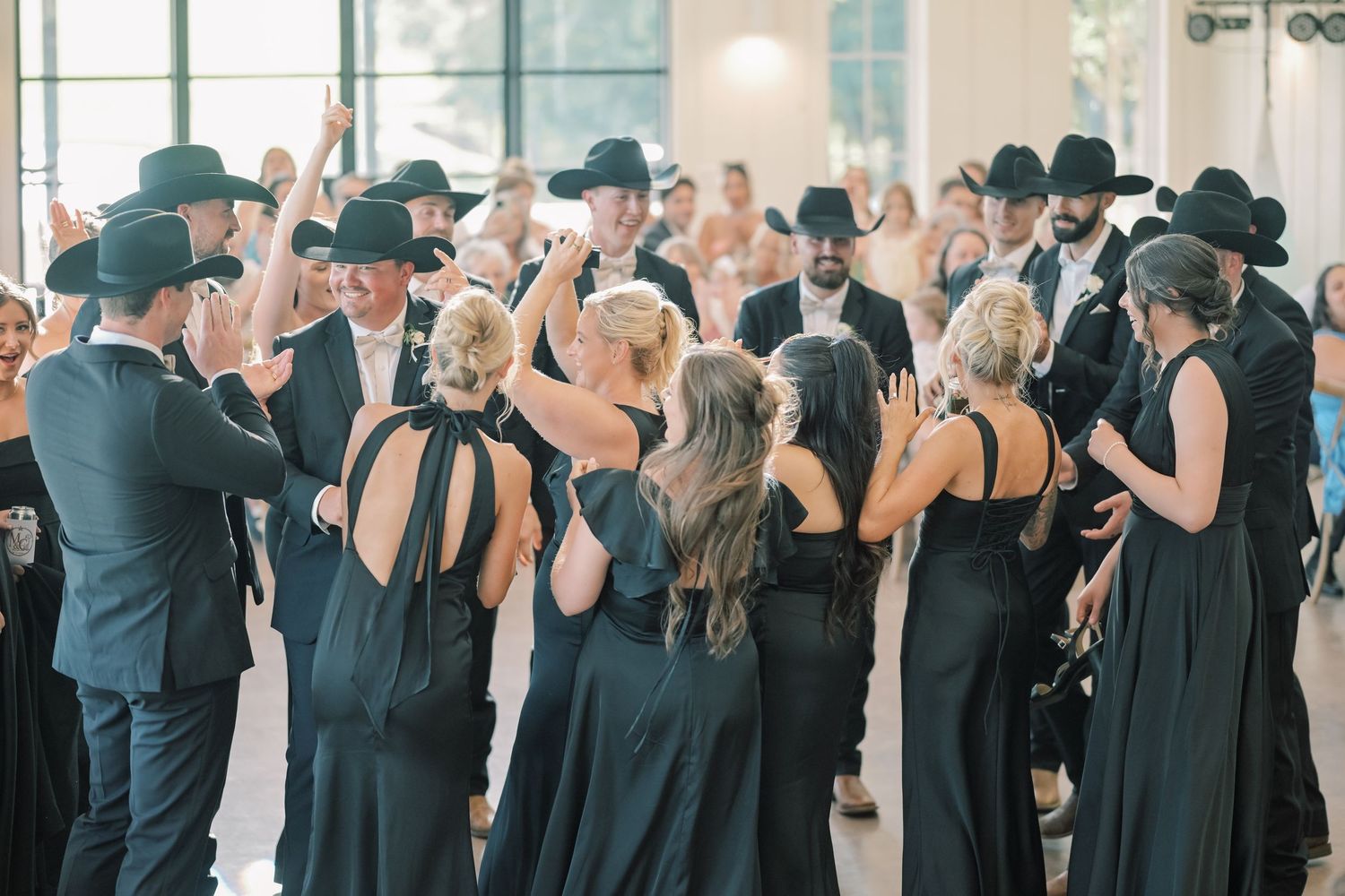 Wedding party in black attire and cowboy hats dance together at indoor reception.