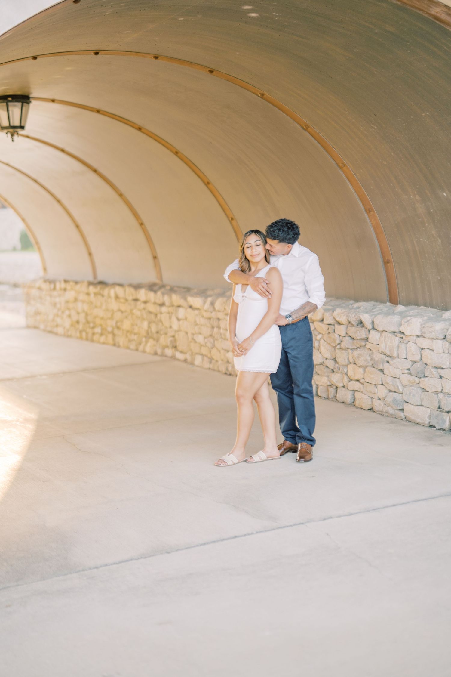 A couple embraces under a curved architectural tunnel, the man kissing the womans shoulder tenderly.