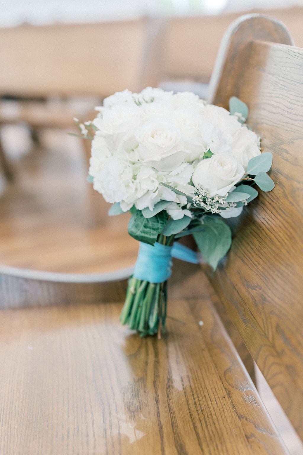 White rose and hydrangea bridal bouquet tied with teal ribbon resting on wooden church pew.