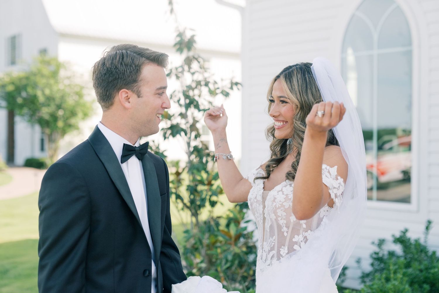 Bride playfully lifts veil while laughing with groom in black tuxedo outside white chapel.