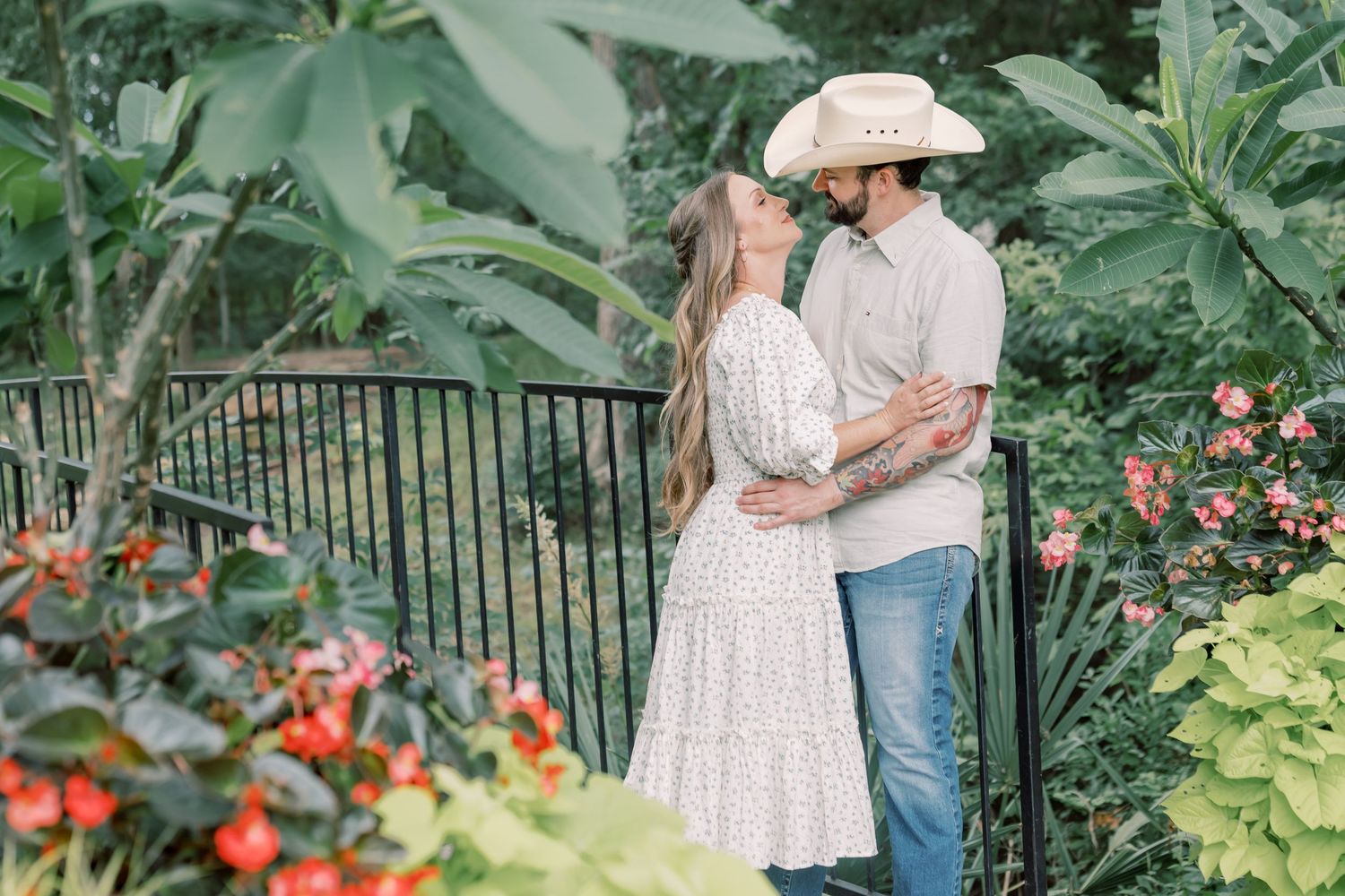 Couple embracing and almost kissing in a lush garden, man wearing cowboy hat, woman in white floral dress.