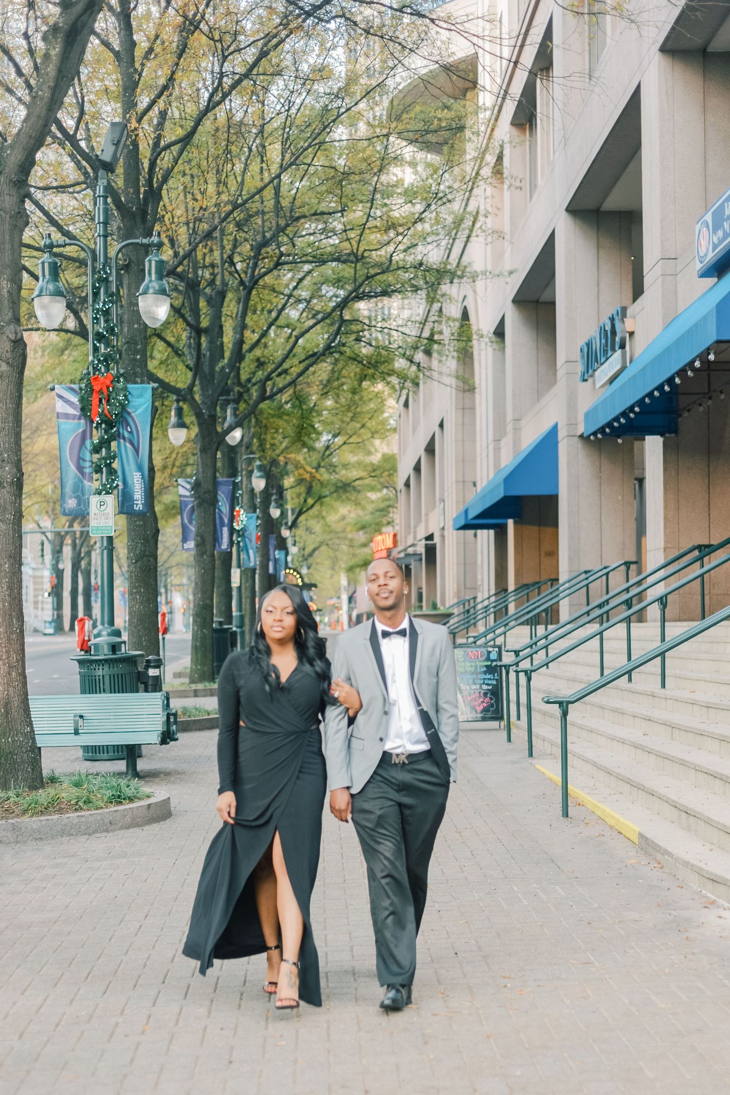 Stylish couple in formal attire walking arm-in-arm along a tree-lined urban sidewalk.