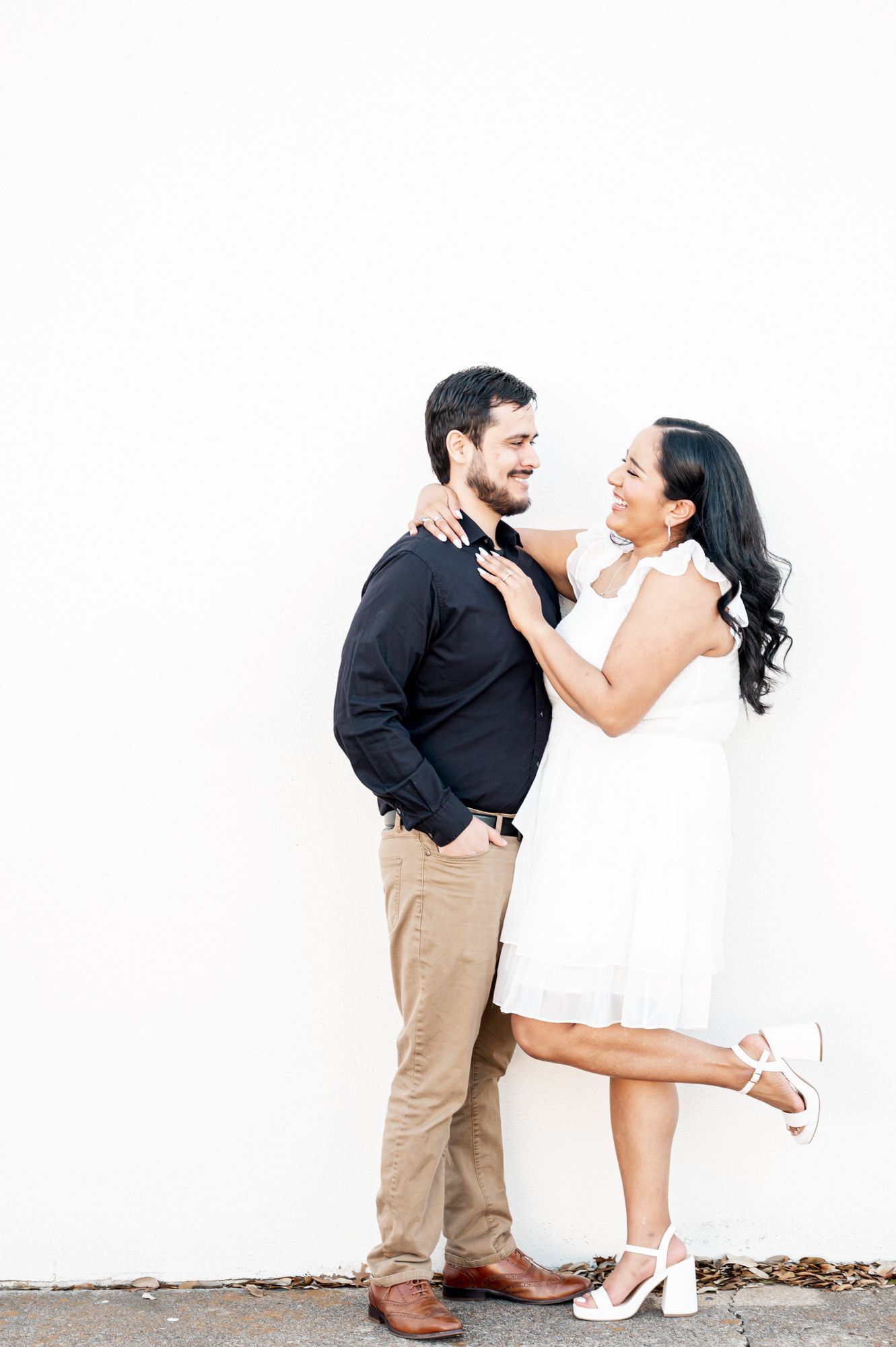 A playful couple in formal attire poses together against a white wall in an intimate engagement photo.