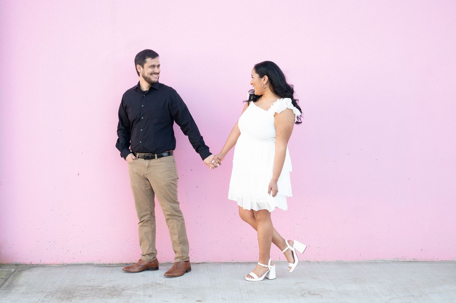 A couple in coordinated outfits smiling and holding hands against a bright pink wall background.