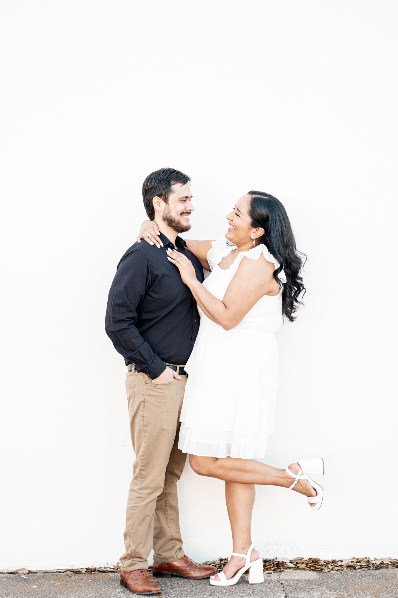Playful couple in white dress and khaki pants pose against white wall.