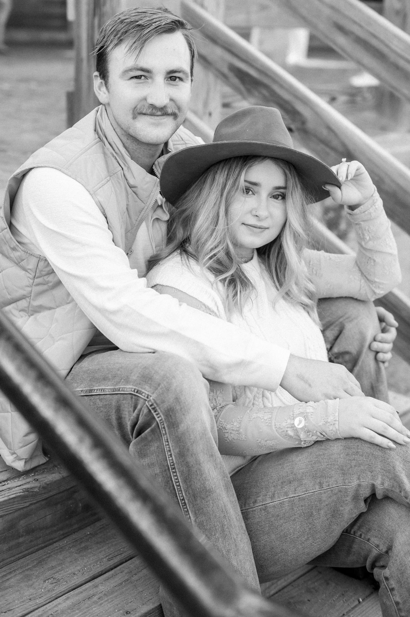 A romantic couple in casual attire shares intimate moments while sitting on wooden steps in black and white photography.