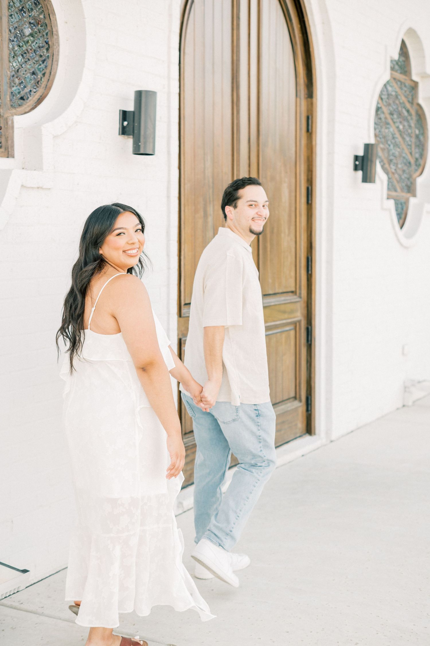 A couple shares a joyful moment walking along a white Mediterranean-style hallway with wooden doors.