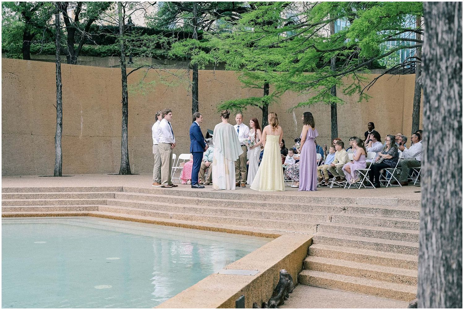 Outdoor wedding ceremony taking place on tiered stone steps beside a tranquil pool surrounded by trees and greenery.