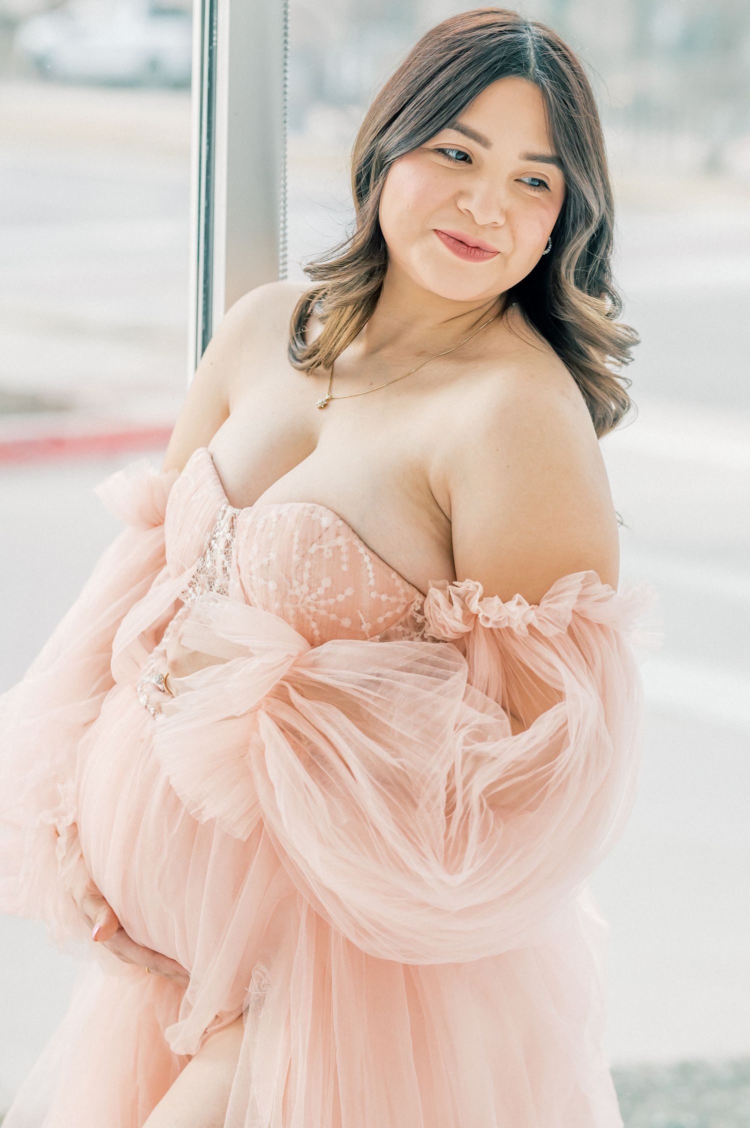 Pregnant woman in blush pink off-shoulder tulle maternity gown posing elegantly near a window.