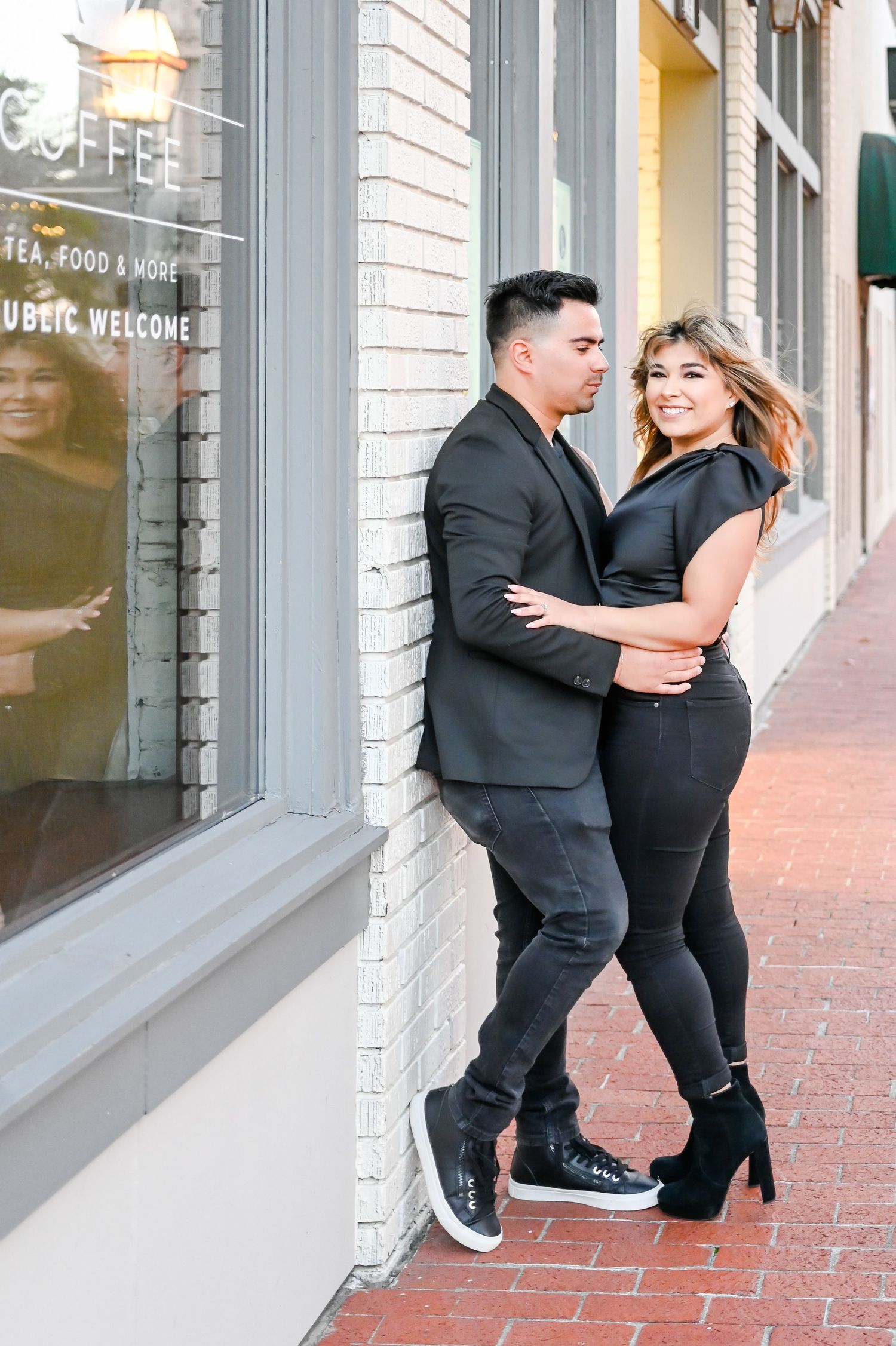 Stylish couple in all-black outfits embrace against a brick wall outside a coffee shop.