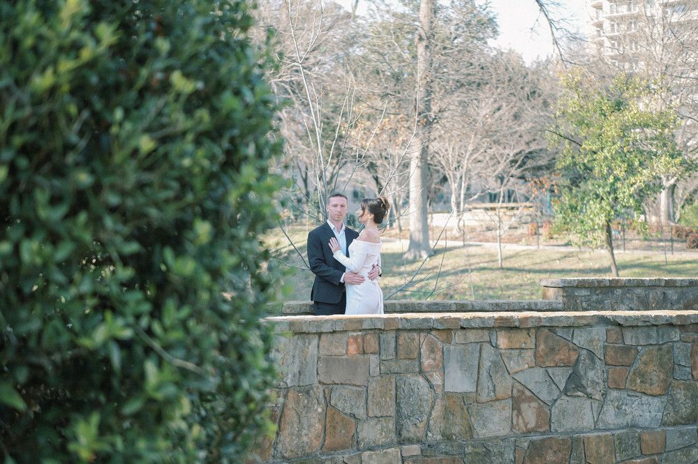 Bride and groom sharing a romantic moment together near a stone wall surrounded by lush garden trees.