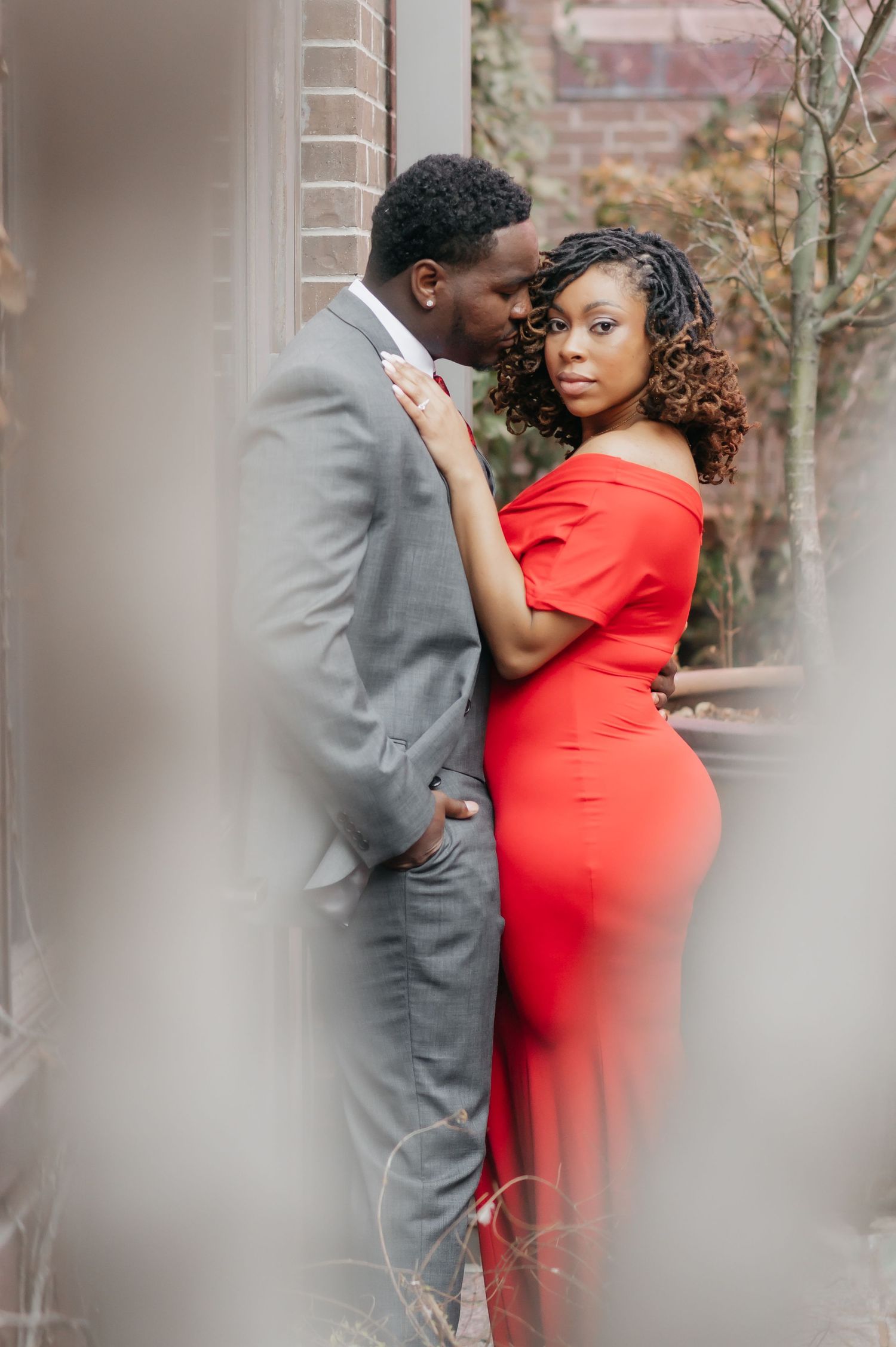Couple in elegant red gown and gray suit share romantic pose against brick wall in garden setting.