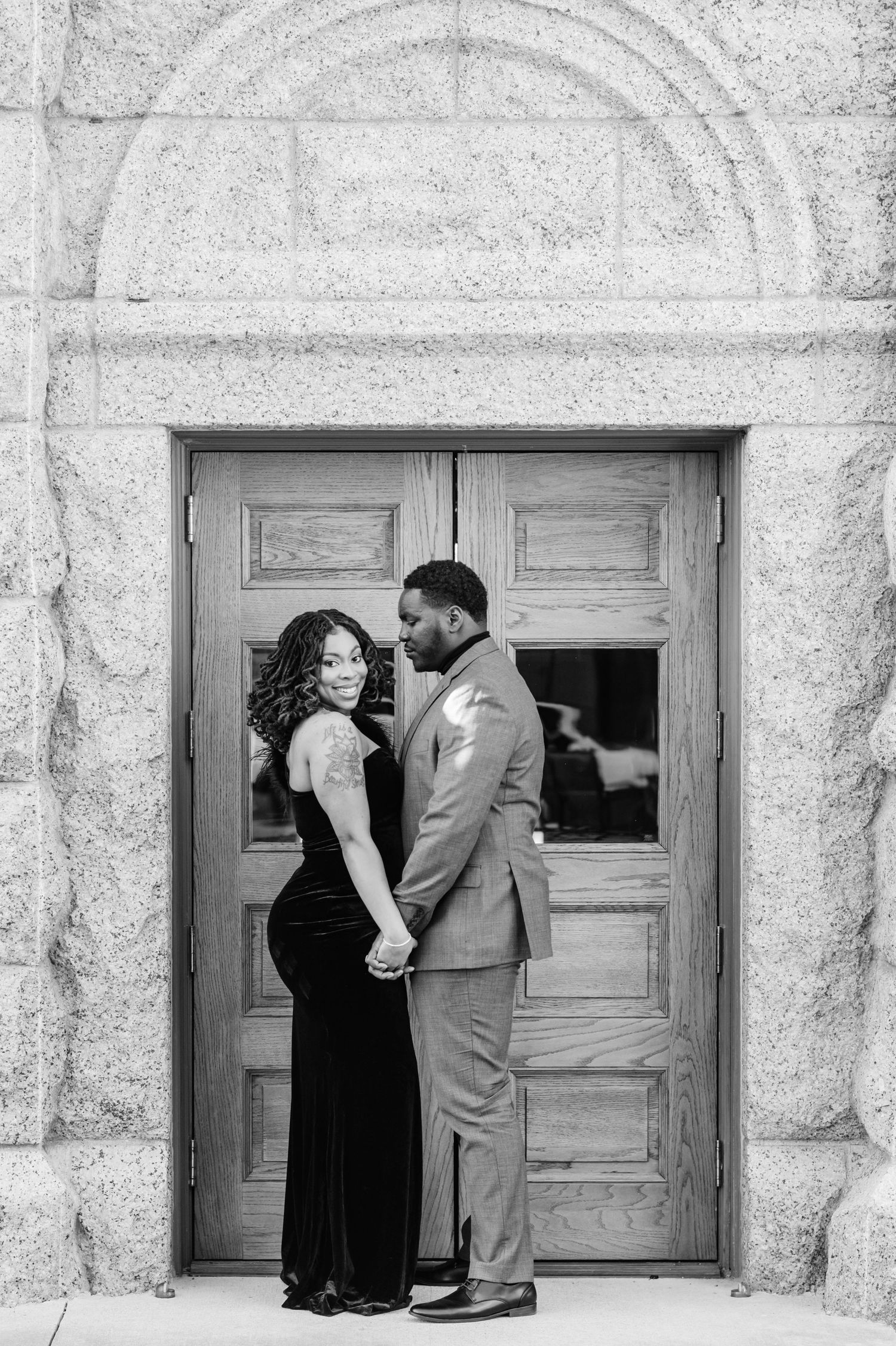 Black and white photo of a couple holding hands and gazing at each other in front of elegant stone building doors.