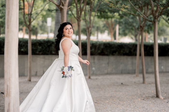 Smiling bride in a flowing white ball gown holding a bouquet among trees outdoors.