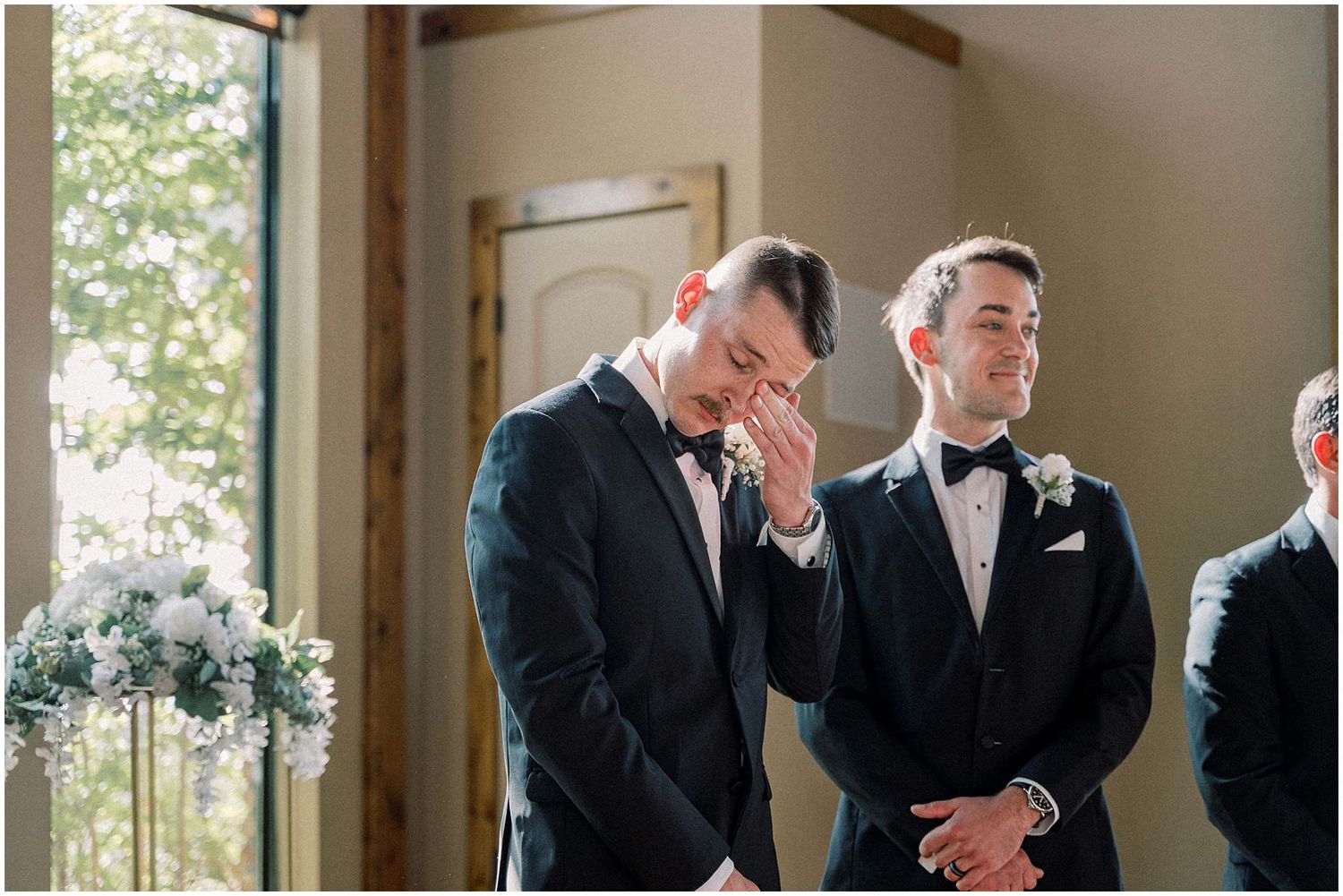 Groomsmen gather in formal attire inside a sunlit room before the ceremony.