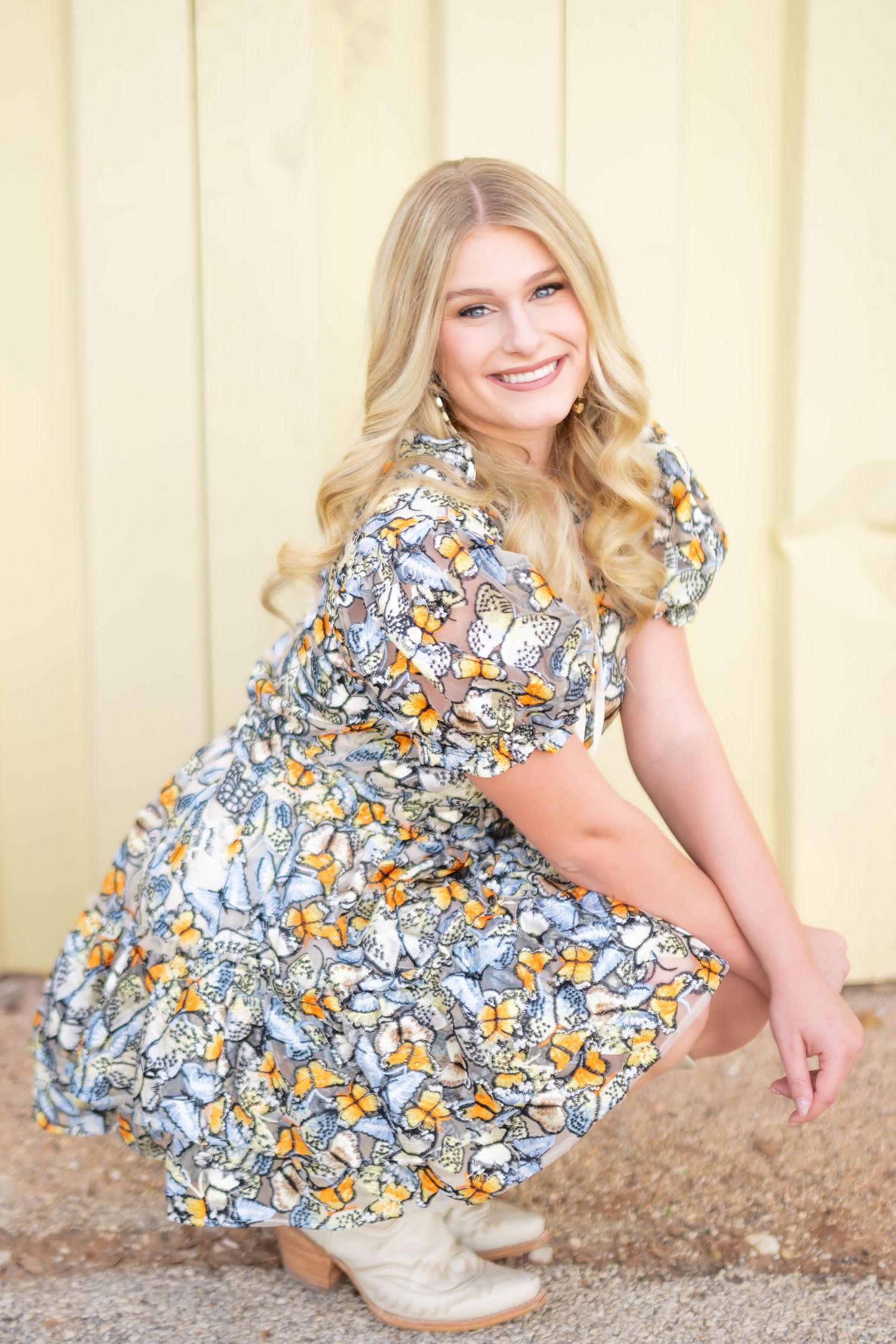 Smiling blonde woman crouching in a floral butterfly print dress and cowboy boots against a yellow wall.