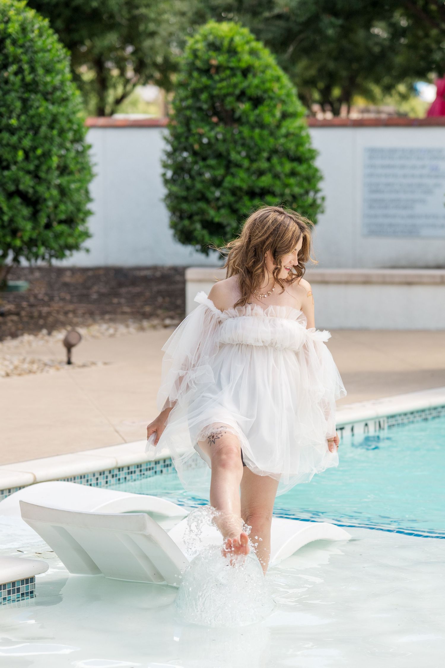 Young girl in white dress splashing in a pool, surrounded by lush green topiaries on a sunny day.