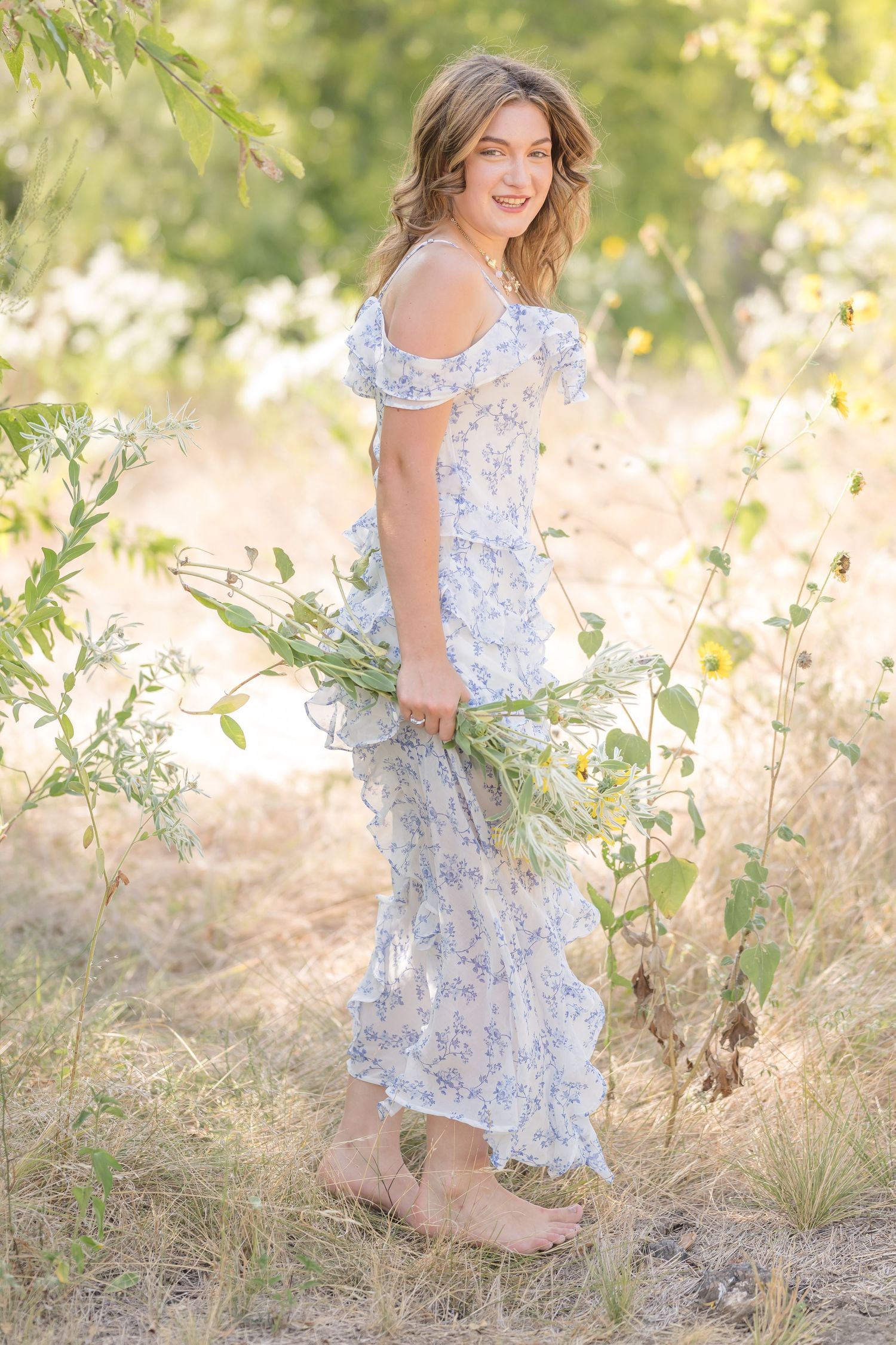 Young woman in floral midi dress holding wildflowers, smiling outdoors in sunlit nature setting.