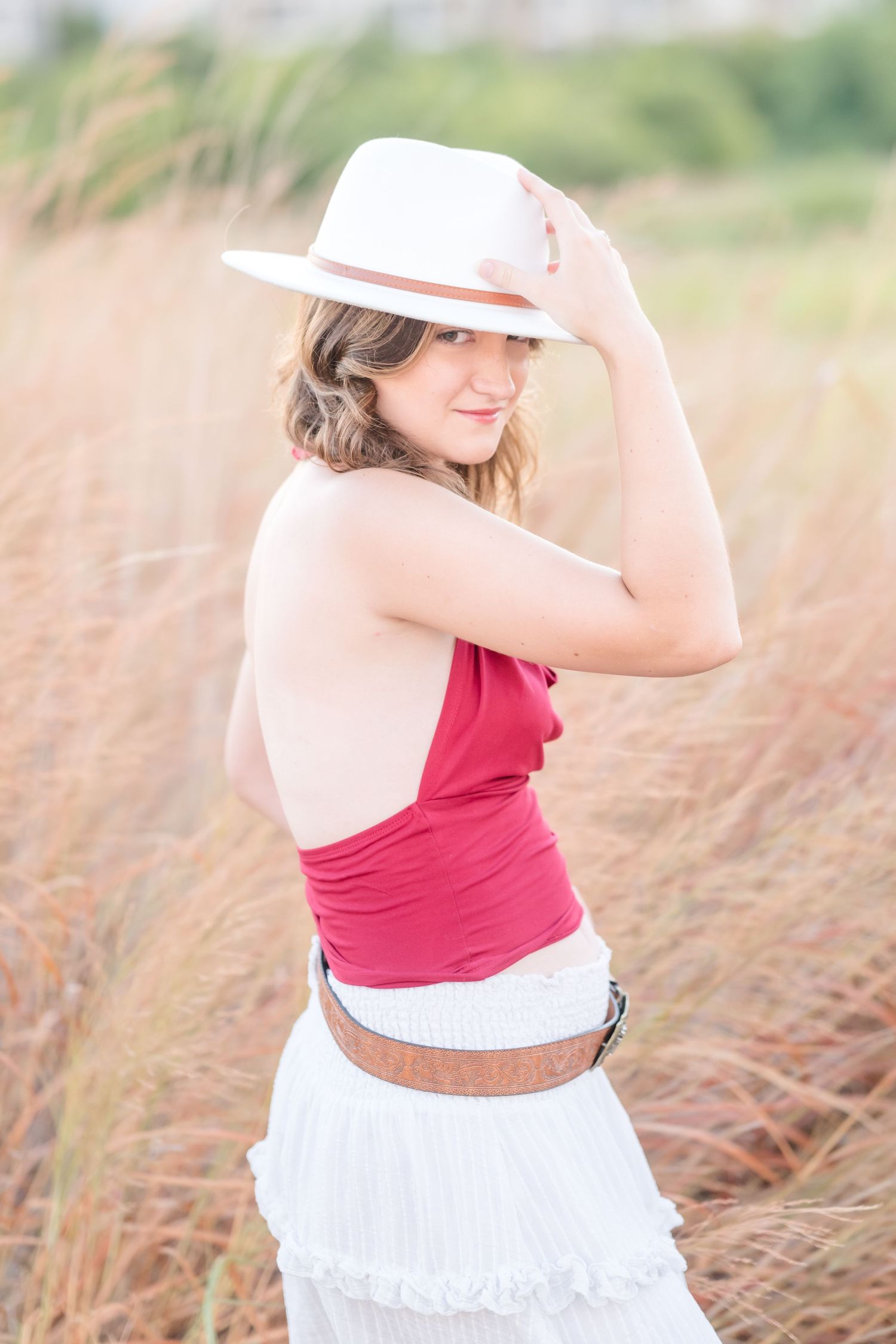 Young woman in red halter top and white hat poses in golden field, smiling over shoulder in stylish summer outfit.