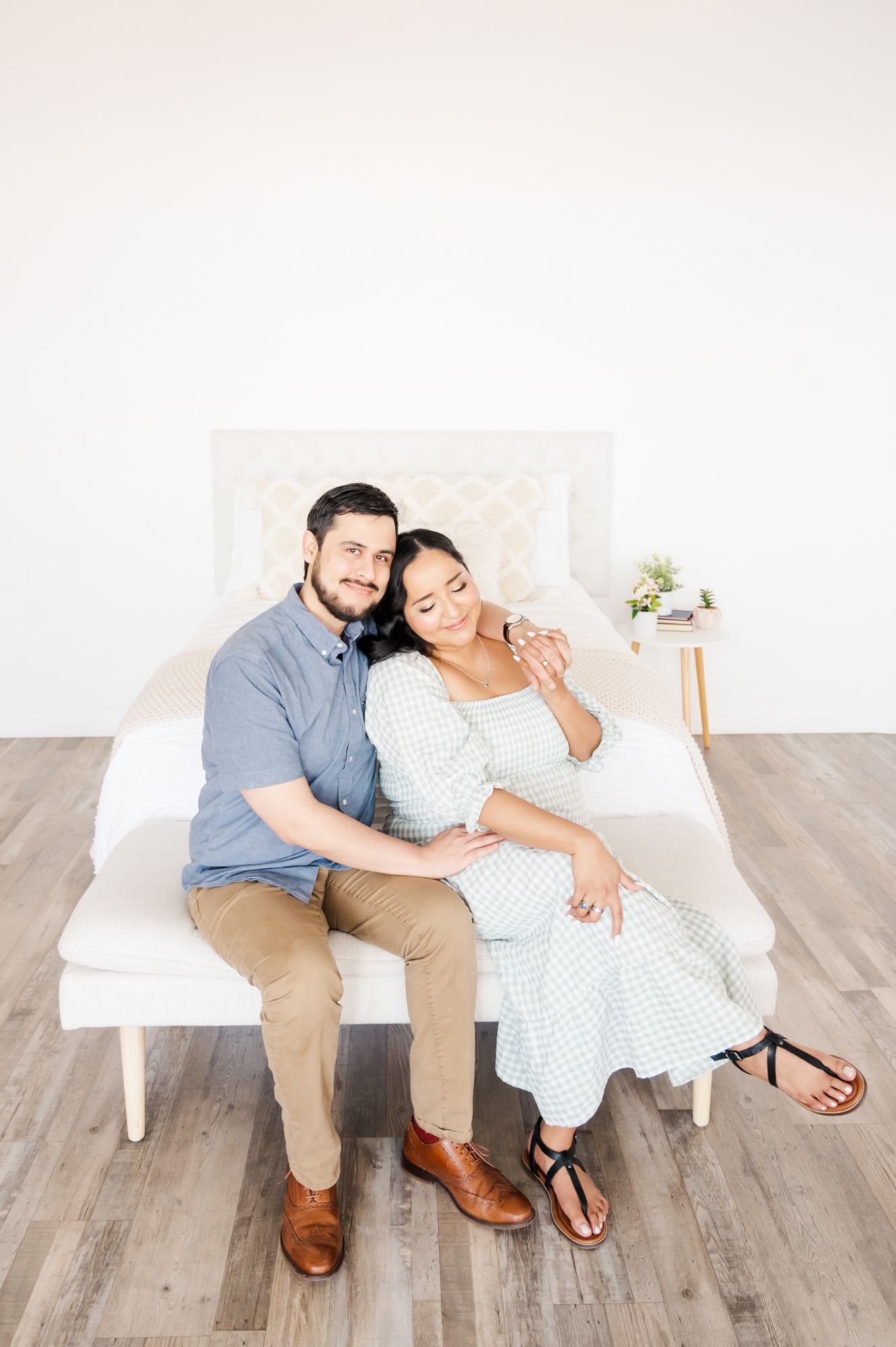 A couple sits together intimately on a white couch against a bright minimal backdrop.