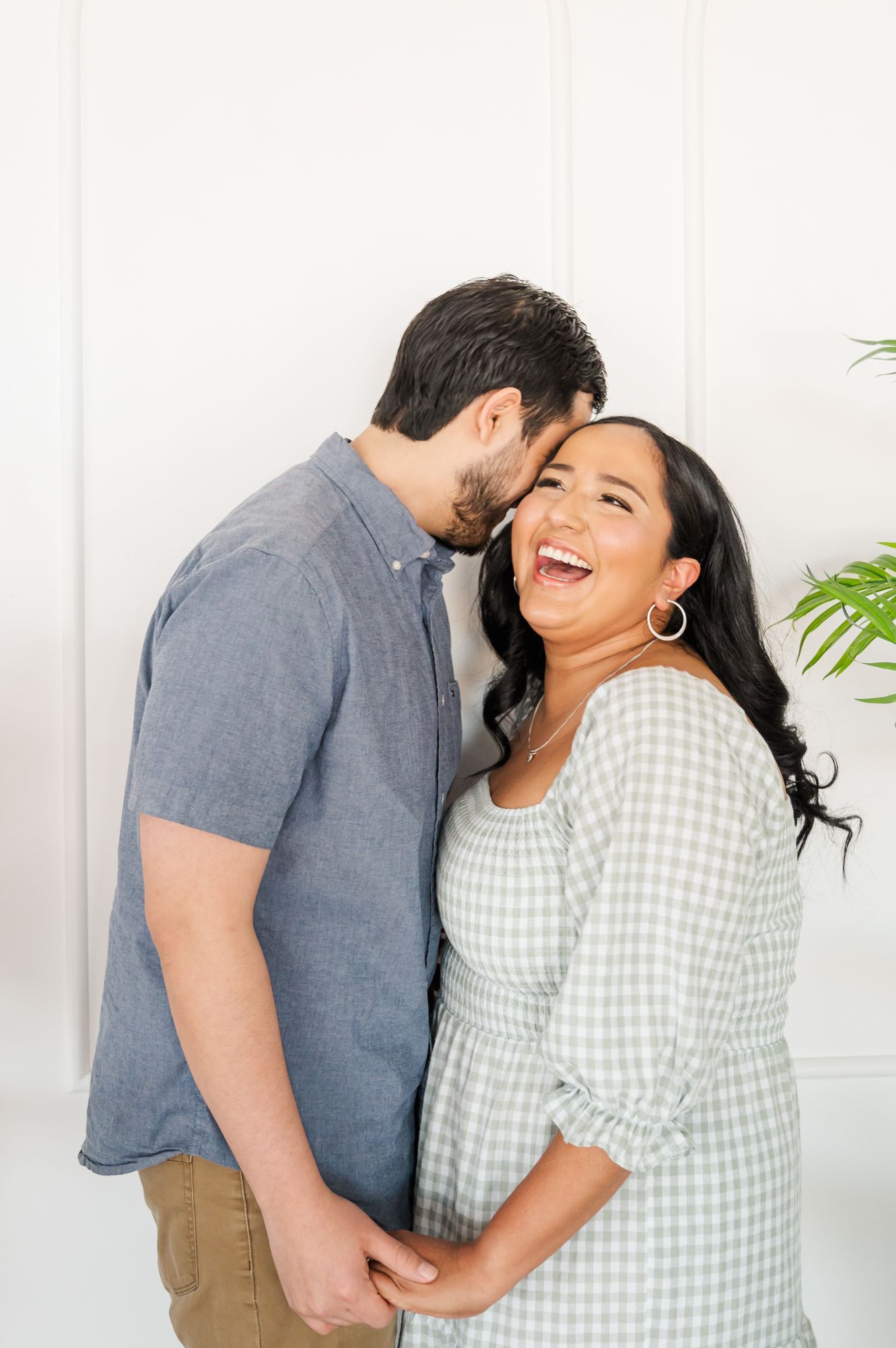 A couple in casual attire sharing intimate moments against a white wall with a green plant in the background.