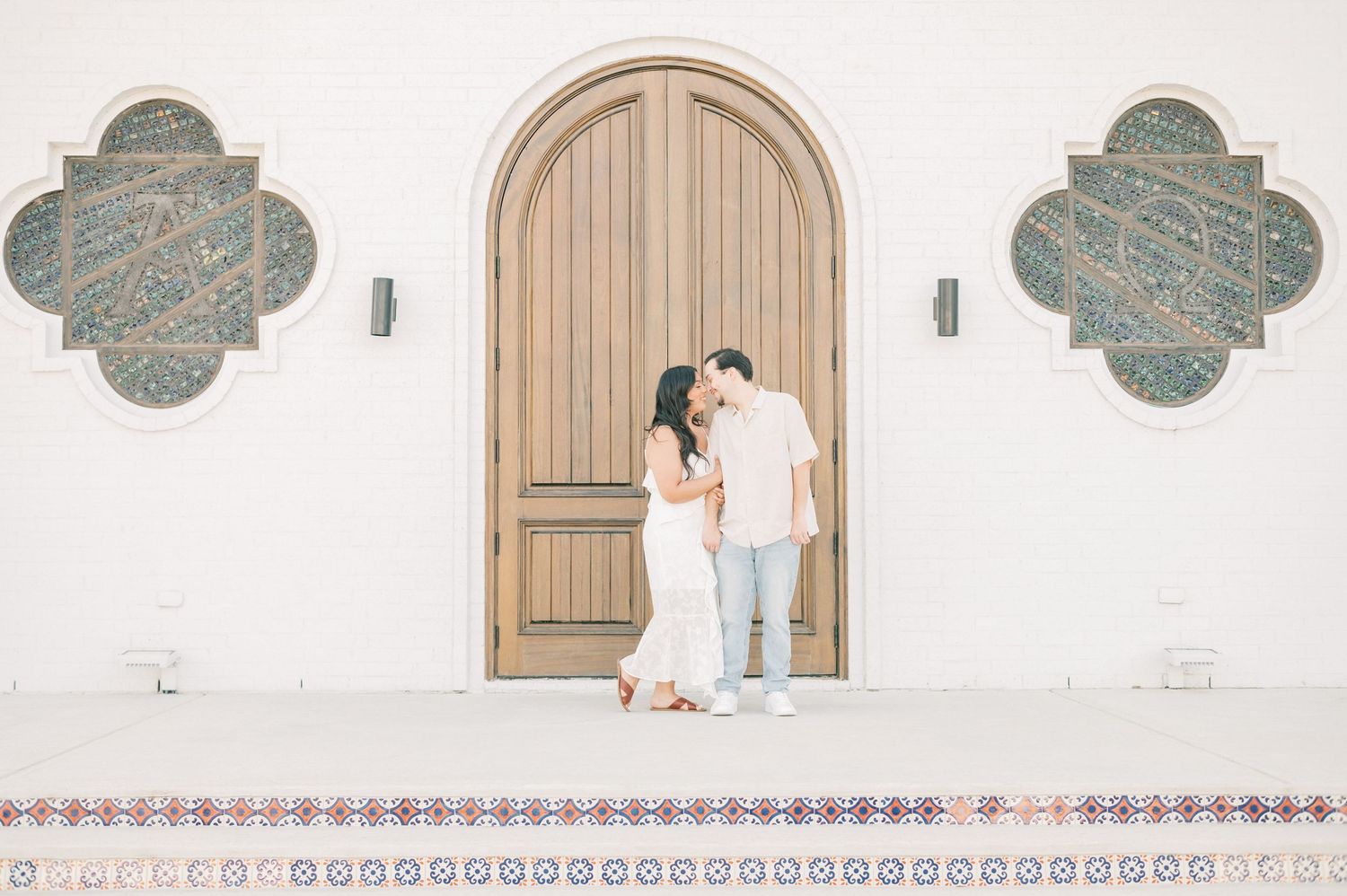 A couple stands intimately in front of an arched wooden door with decorative quatrefoil windows on a white church exterior.