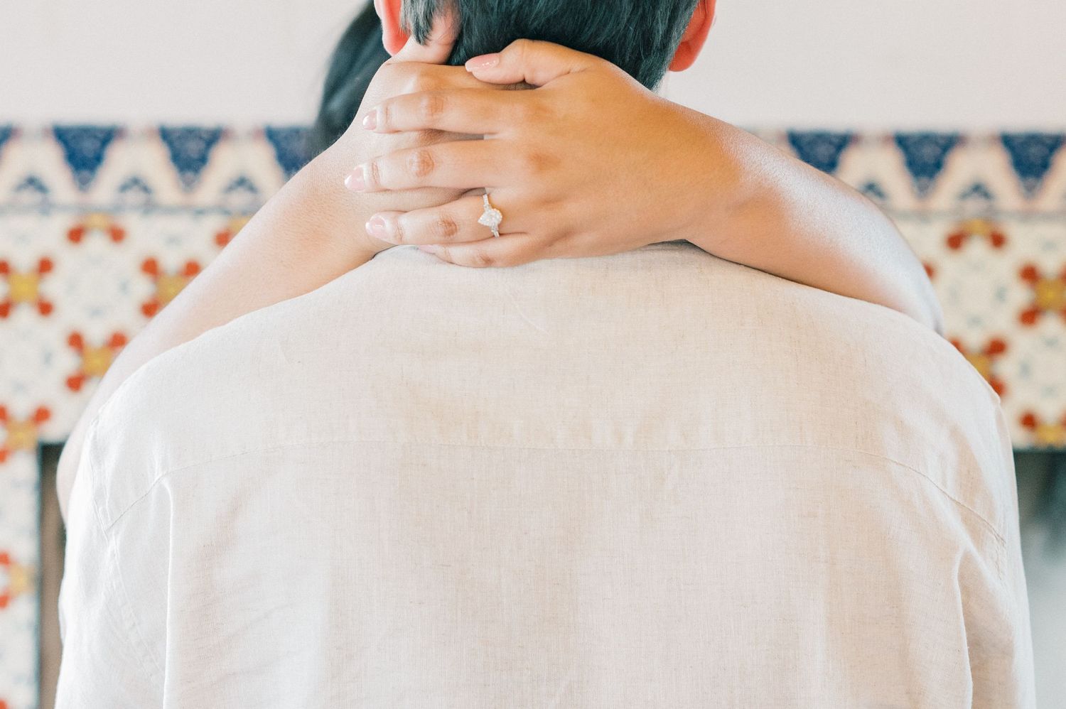 Hands wearing a diamond ring embrace around a white-clothed shoulder against a patterned background.