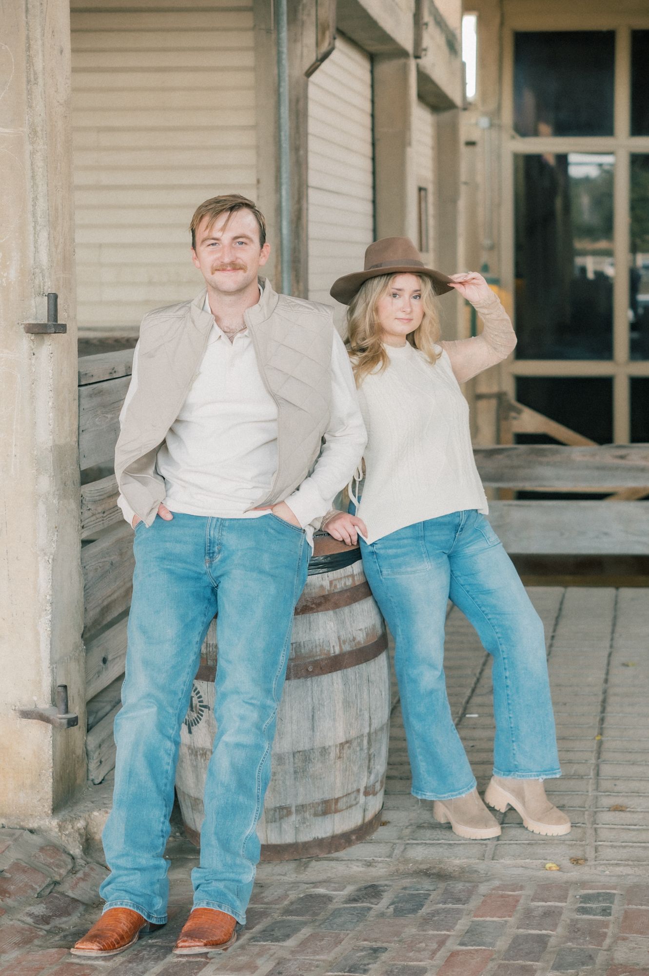 Casual couple in matching denim and light tops posing on wooden porch steps near vintage barrel.