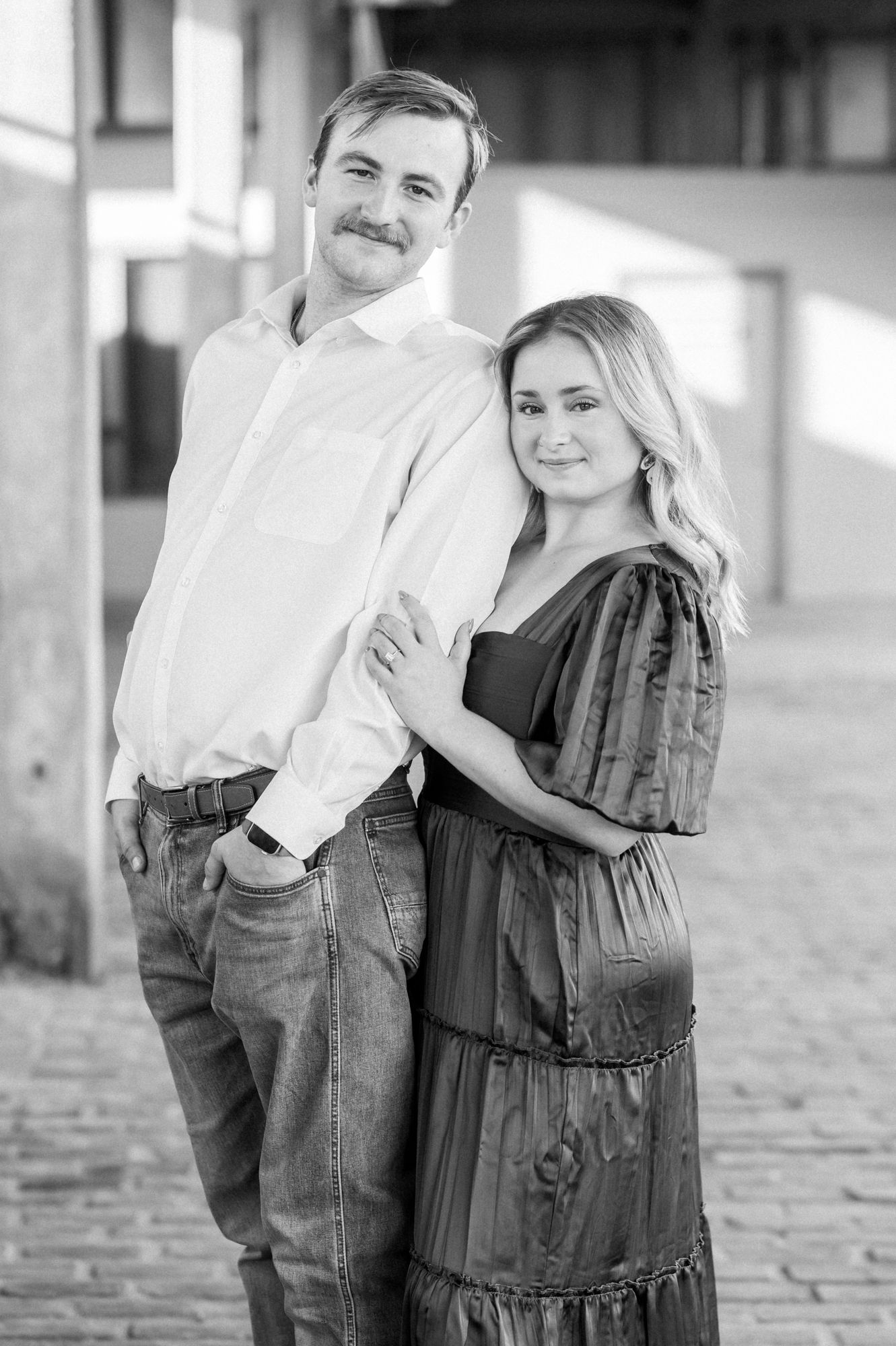A couple in casual attire embraces while standing outdoors on a cobblestone surface in black and white photography.