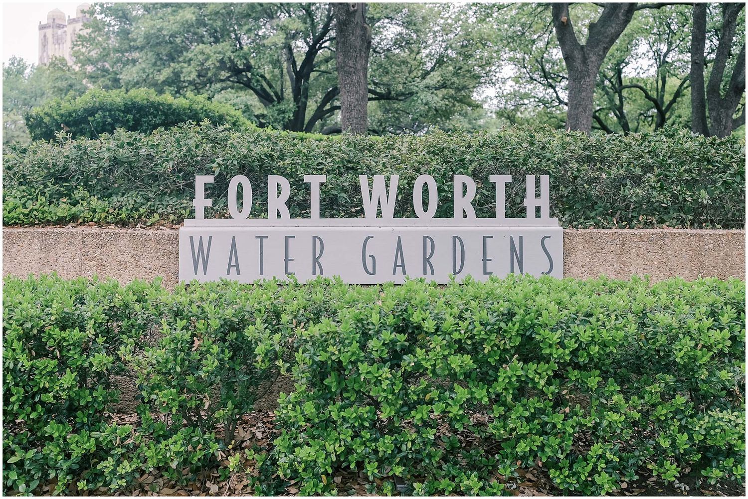 A stone sign for Fort Worth Water Gardens surrounded by lush green shrubs and trees.