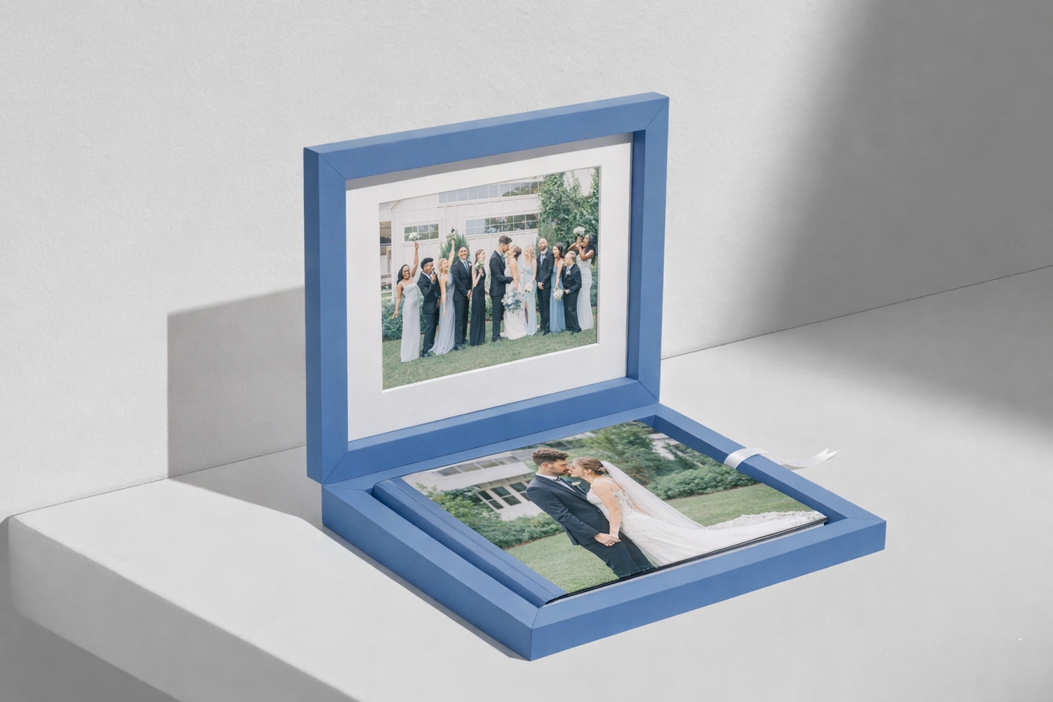 Blue wooden photo box displaying wedding photos, including a framed bridal party print and an open album on a white surface.
