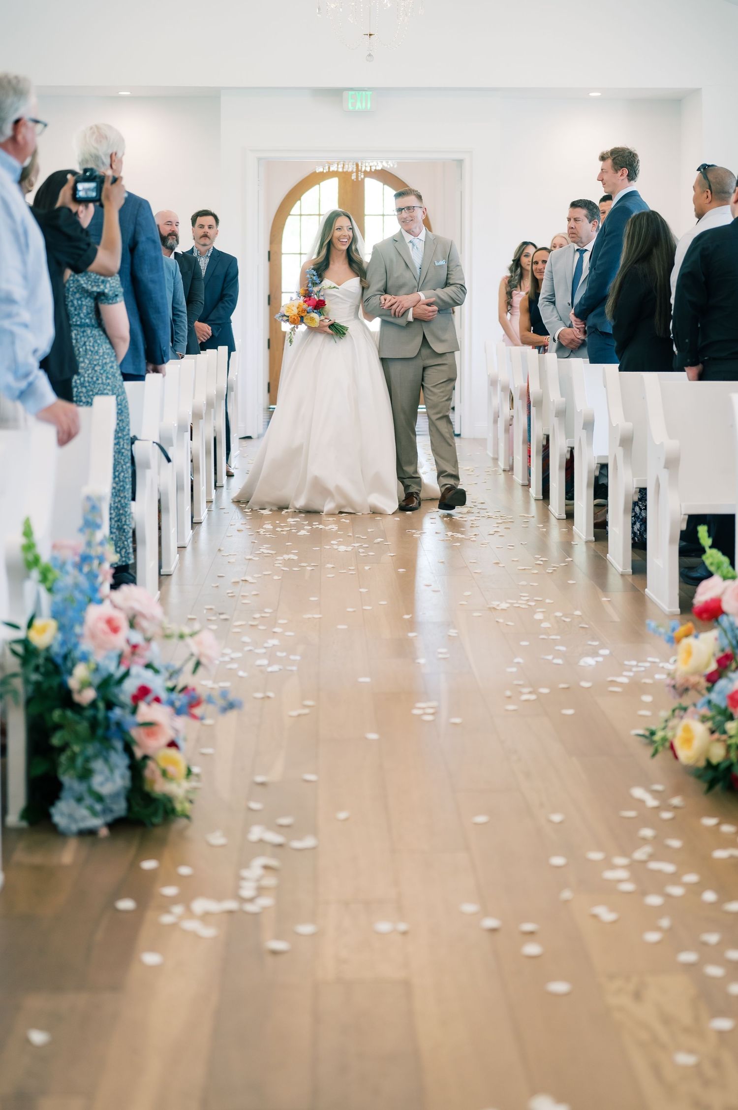 Bride and groom walk down flower petal-lined aisle in bright white chapel surrounded by colorful floral arrangements.