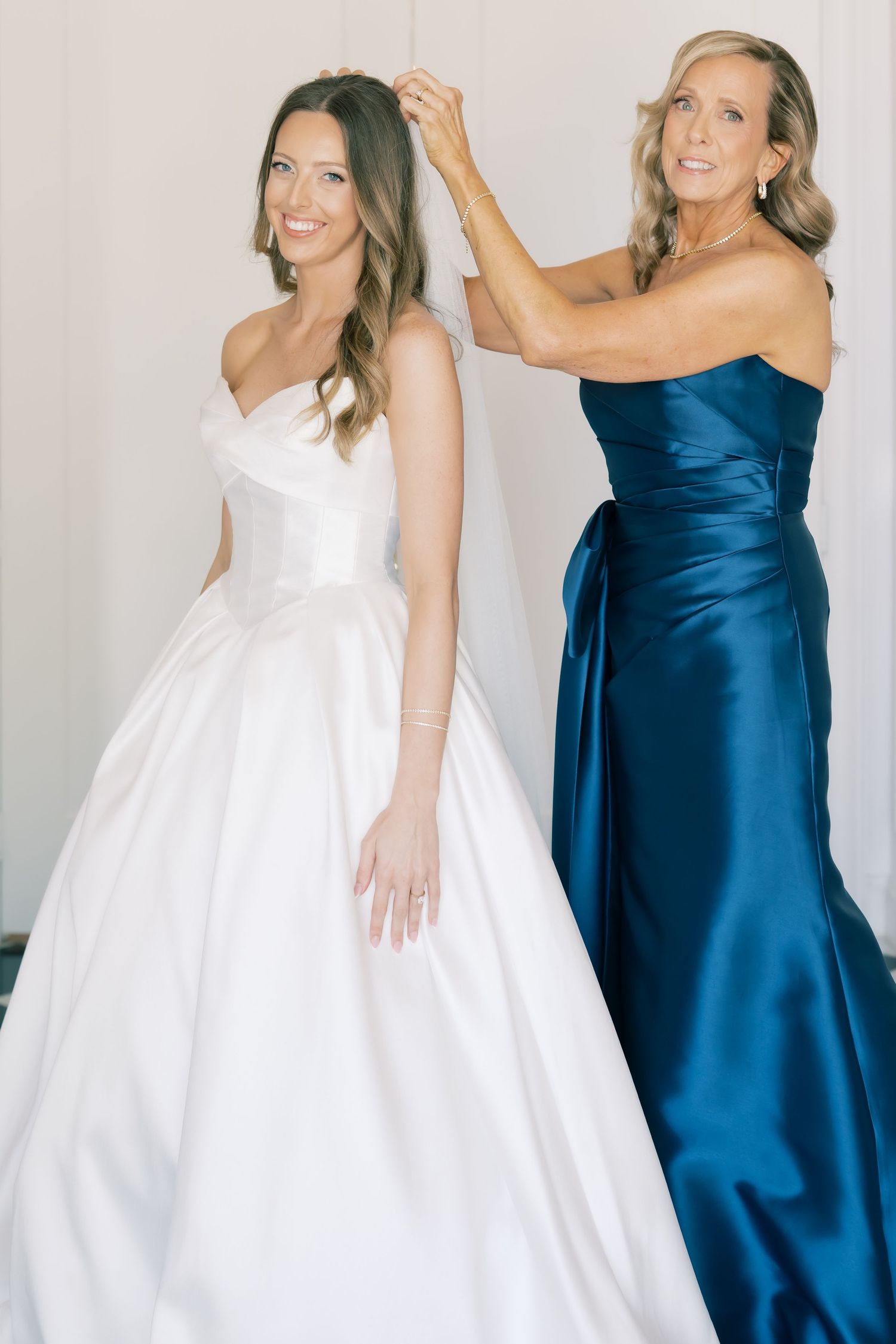 Bride in strapless white ballgown smiles as woman in blue satin dress adjusts her hair.