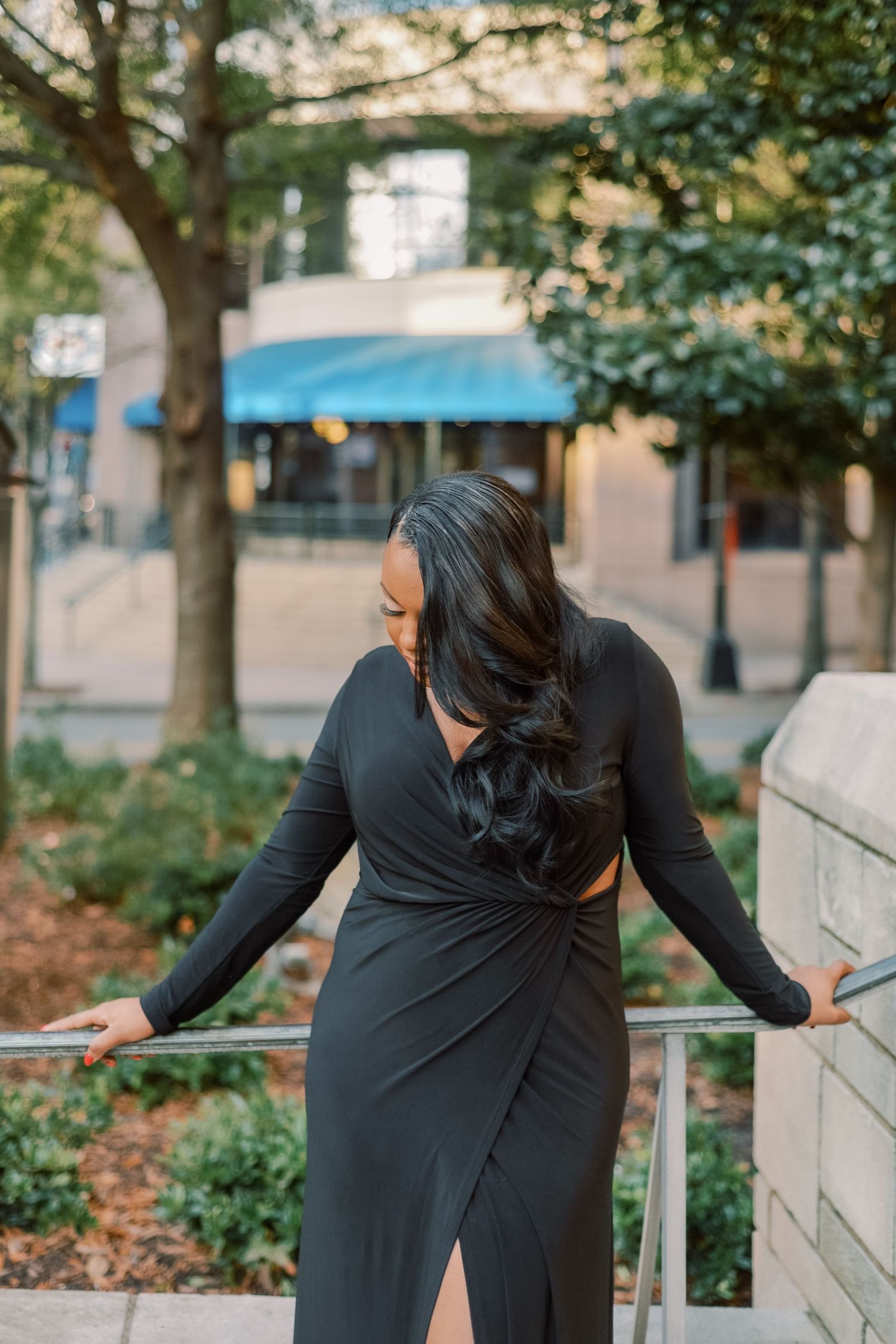 Woman in elegant black wrap dress with open back poses on urban railing surrounded by lush greenery.