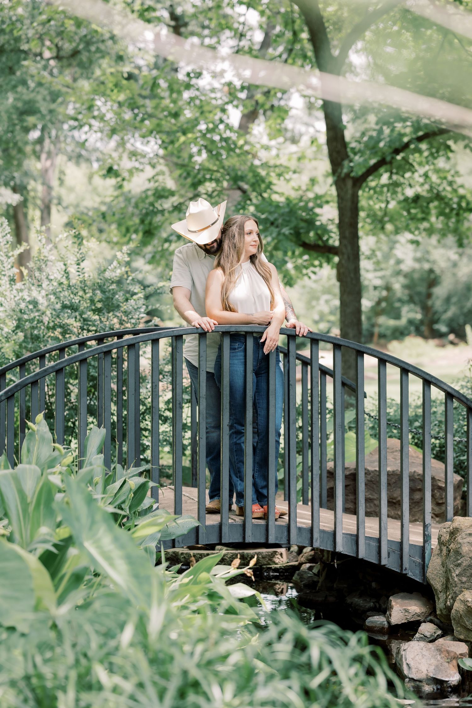 Couple standing on a charming arched bridge surrounded by lush greenery, man in cowboy hat embracing woman in jeans.