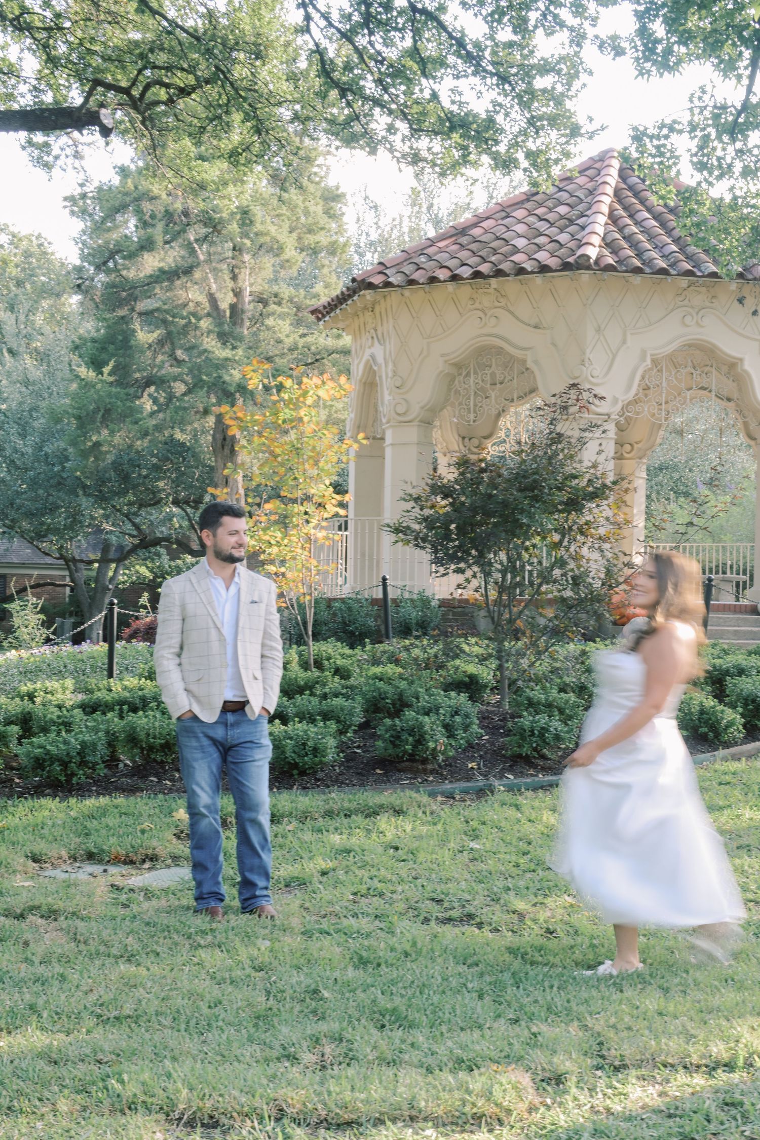 Man in jeans and a blazer stands on the grass as a woman in a white dress twirls near a stone gazebo in a garden engagement shoot.