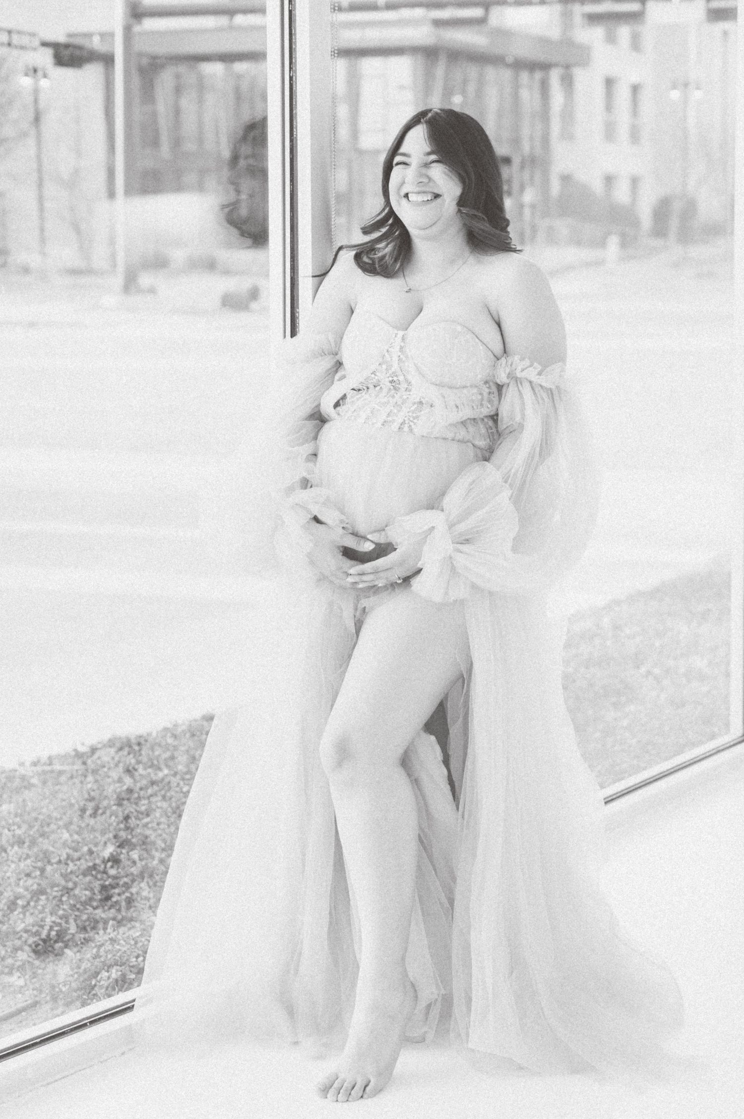 Black and white maternity photo of smiling pregnant woman in flowing tulle gown posing by large window.