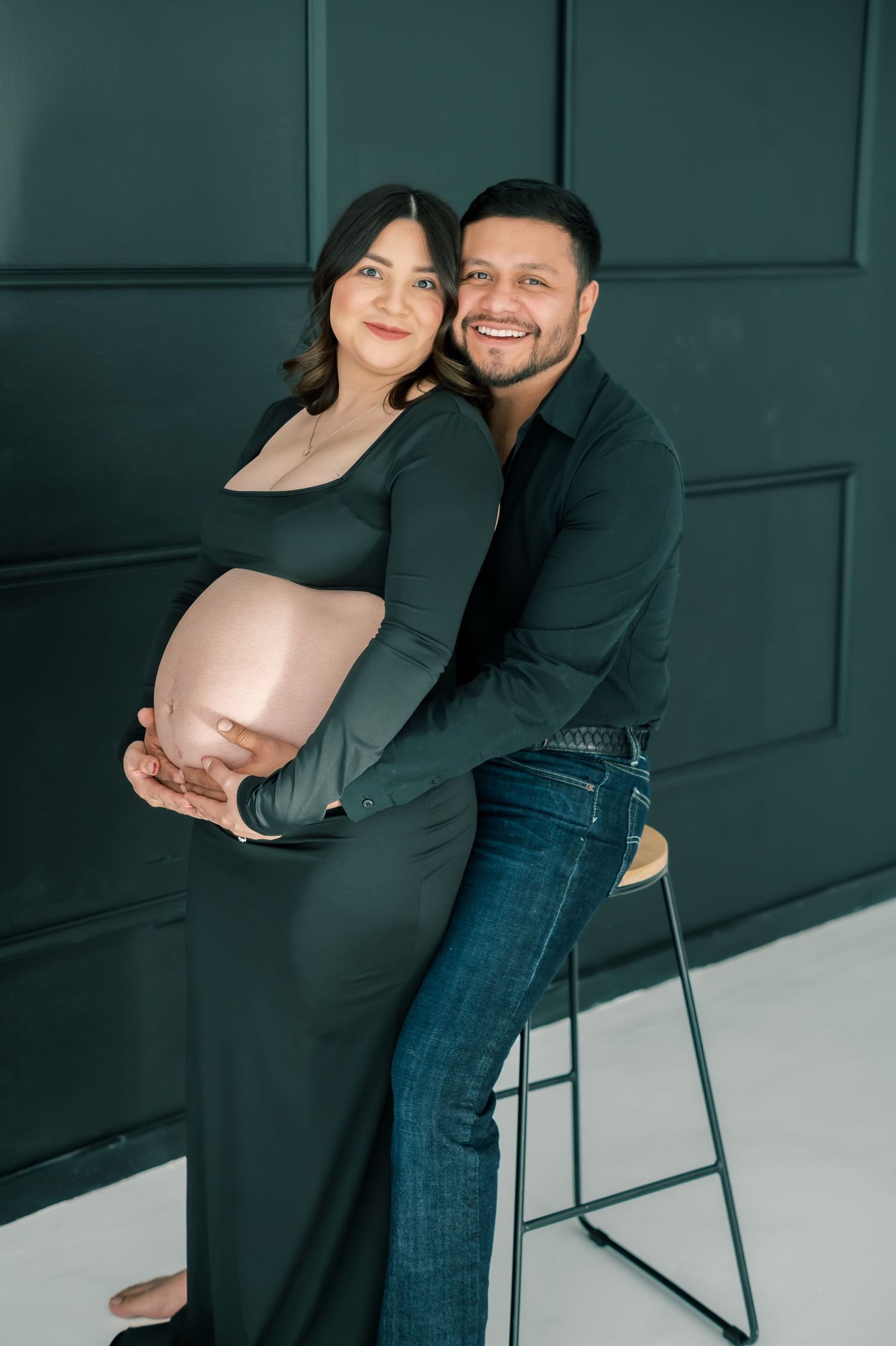 Smiling couple in matching dark green outfits during maternity photo shoot, man embracing pregnant woman from behind.