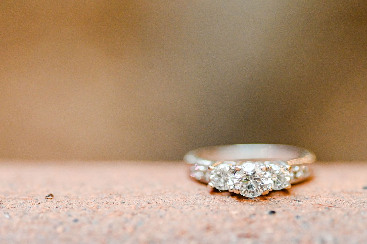 Three-diamond engagement ring in white gold setting displayed on textured surface with soft bokeh background.