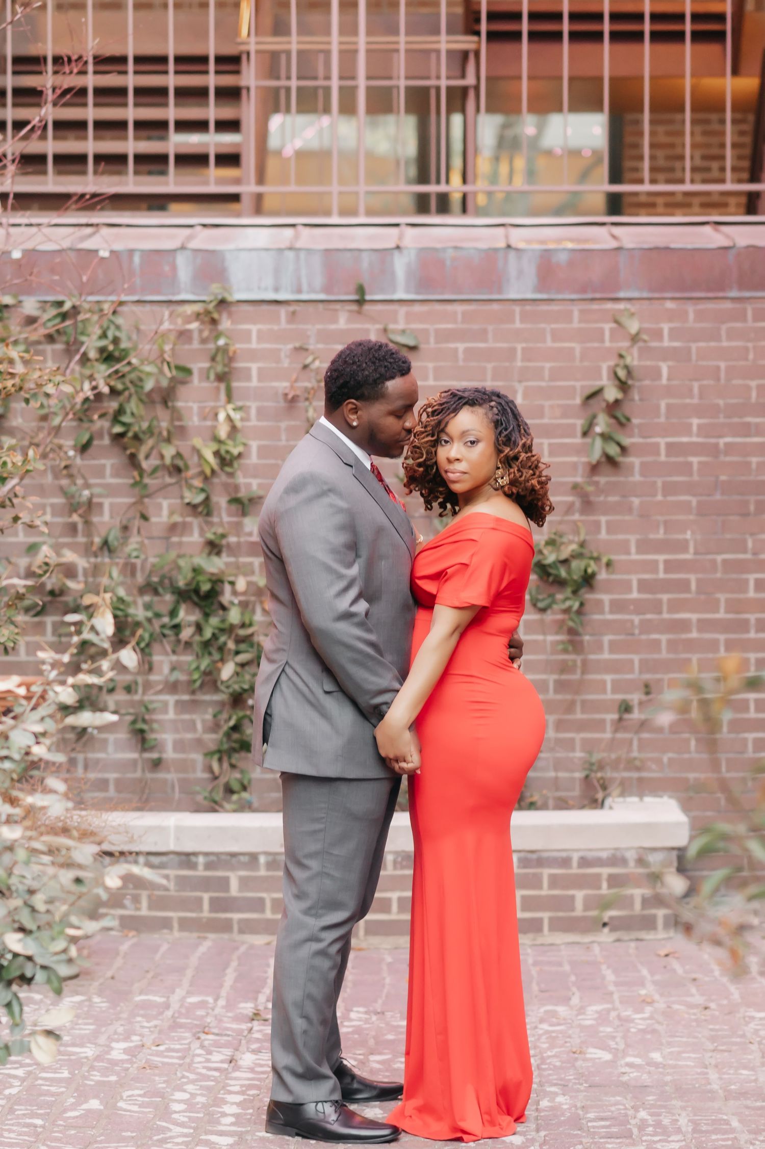 Elegant couple posing against brick wall, woman in red off-shoulder gown, man in gray suit, engagement photo.