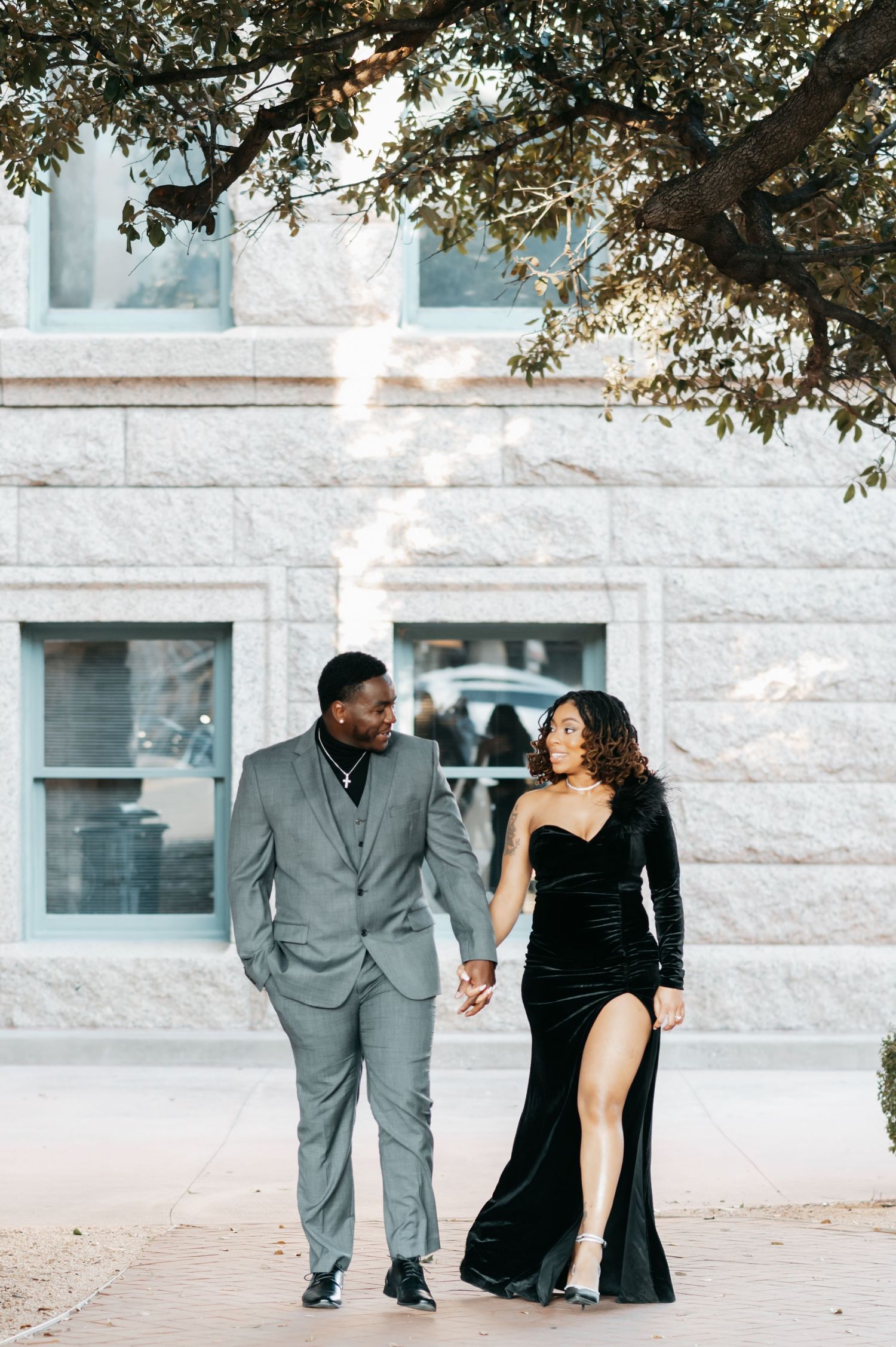 Elegant couple holding hands outdoors, man in gray suit and woman in black velvet one-shoulder gown, posing near stone building.